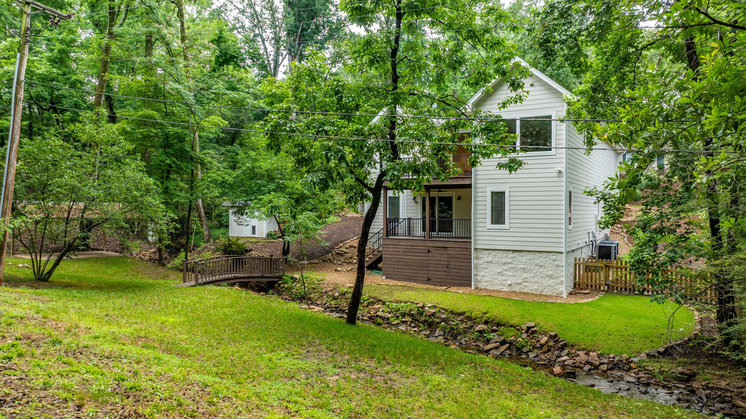 A house in a wooded backyard with a small wooden bridge over a creek, surrounded by green trees and grass.