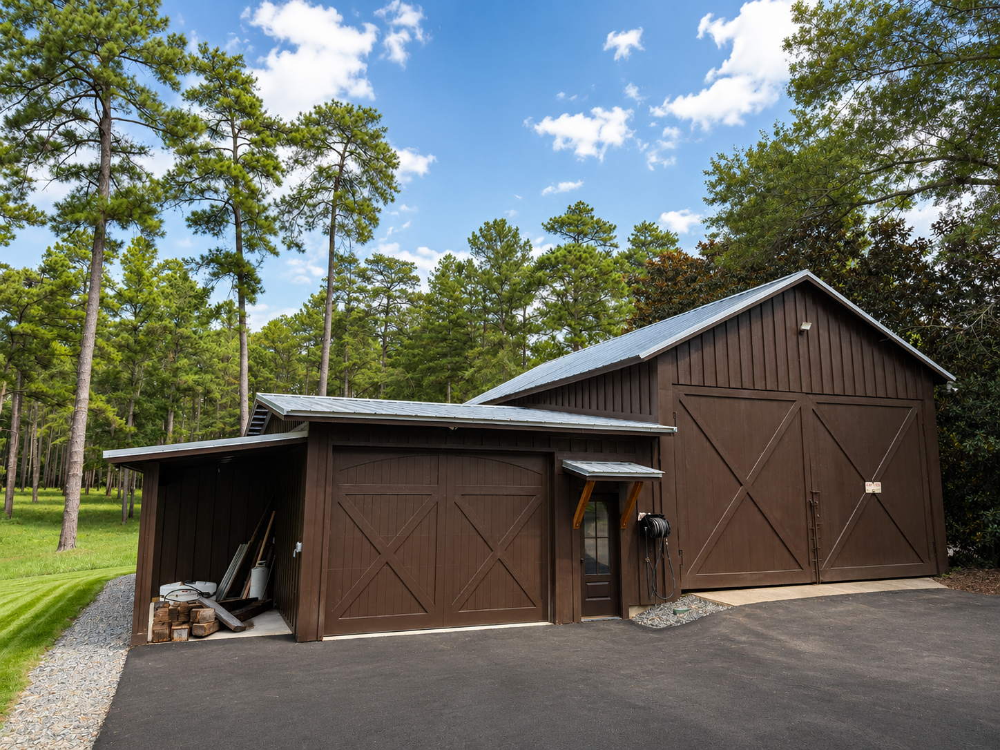A large wooden barn with a metal roof, surrounded by tall pine trees and a bright blue sky with puffy clouds.