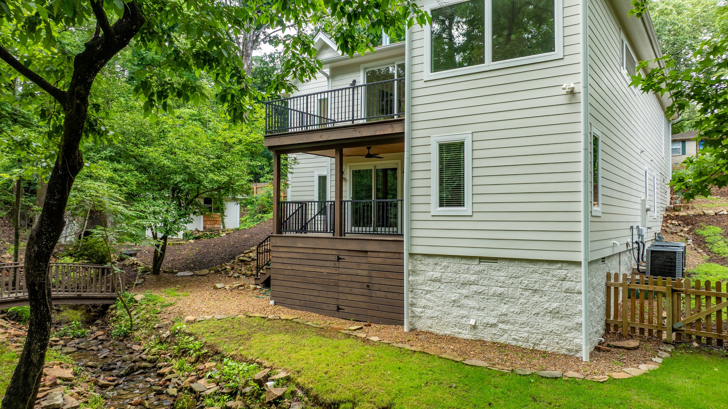Rear view of a two-story house with a small balcony, surrounded by trees and greenery, with a small wooden fence on the right and a landscaped yard.