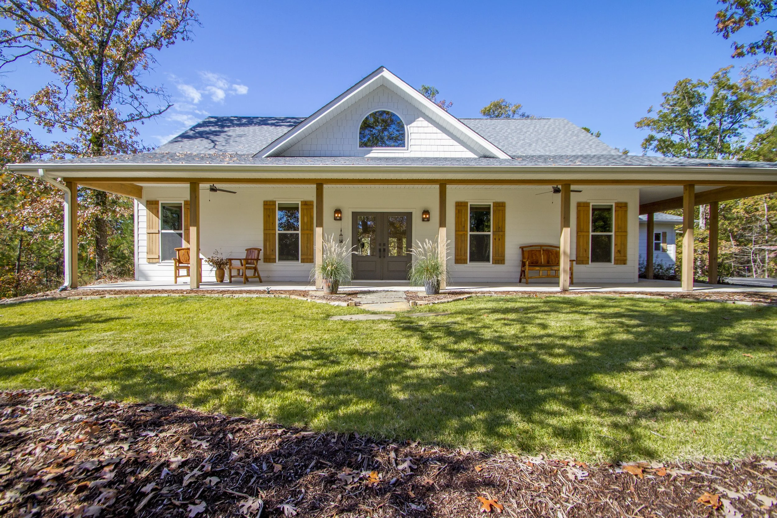 Front view of a white house with a porch, wooden shutters, and a gray roof, surrounded by green grass and trees, under a blue sky.