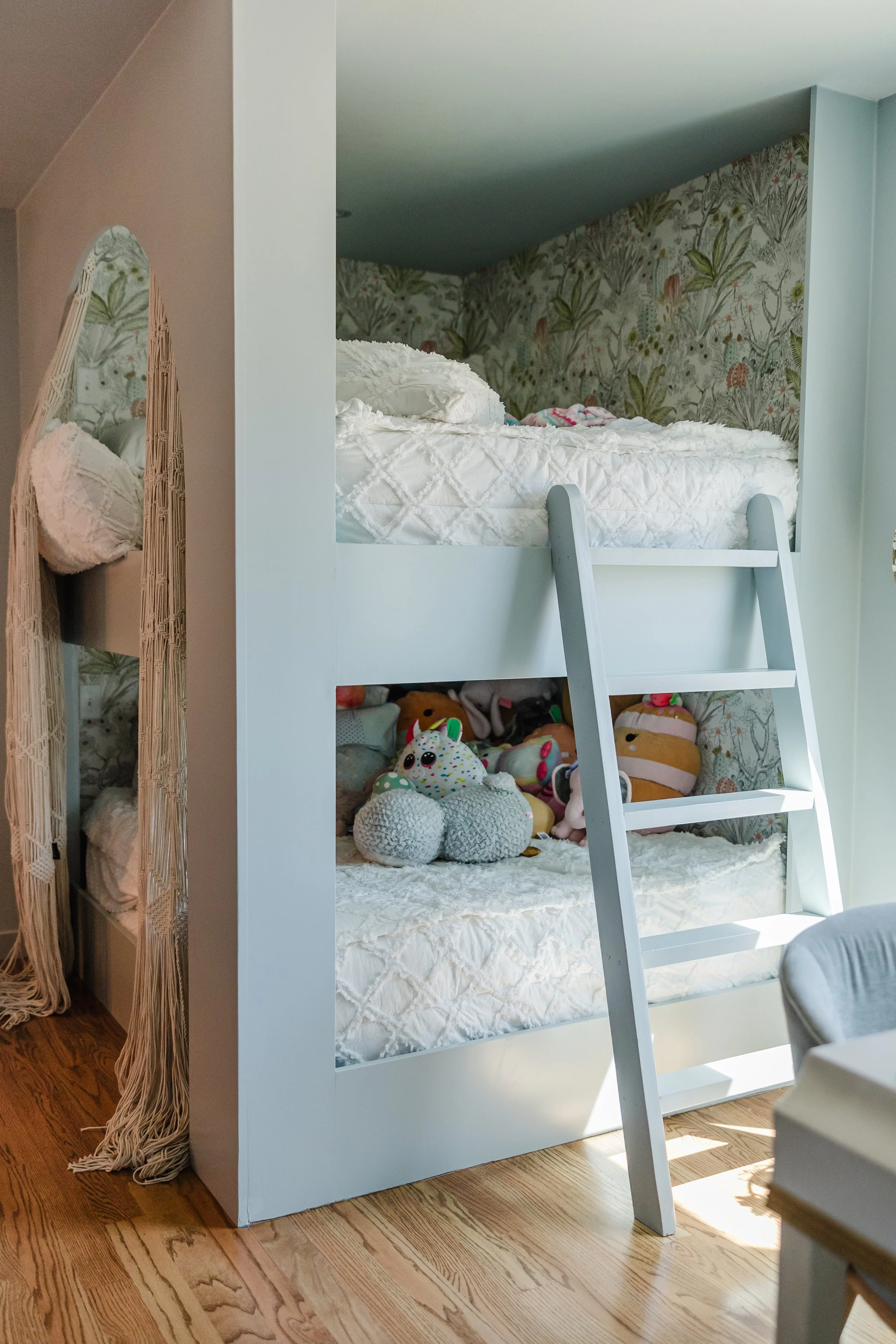 White bunk bed with stuffed animals and plush toys underneath, and floral wallpaper in the background.