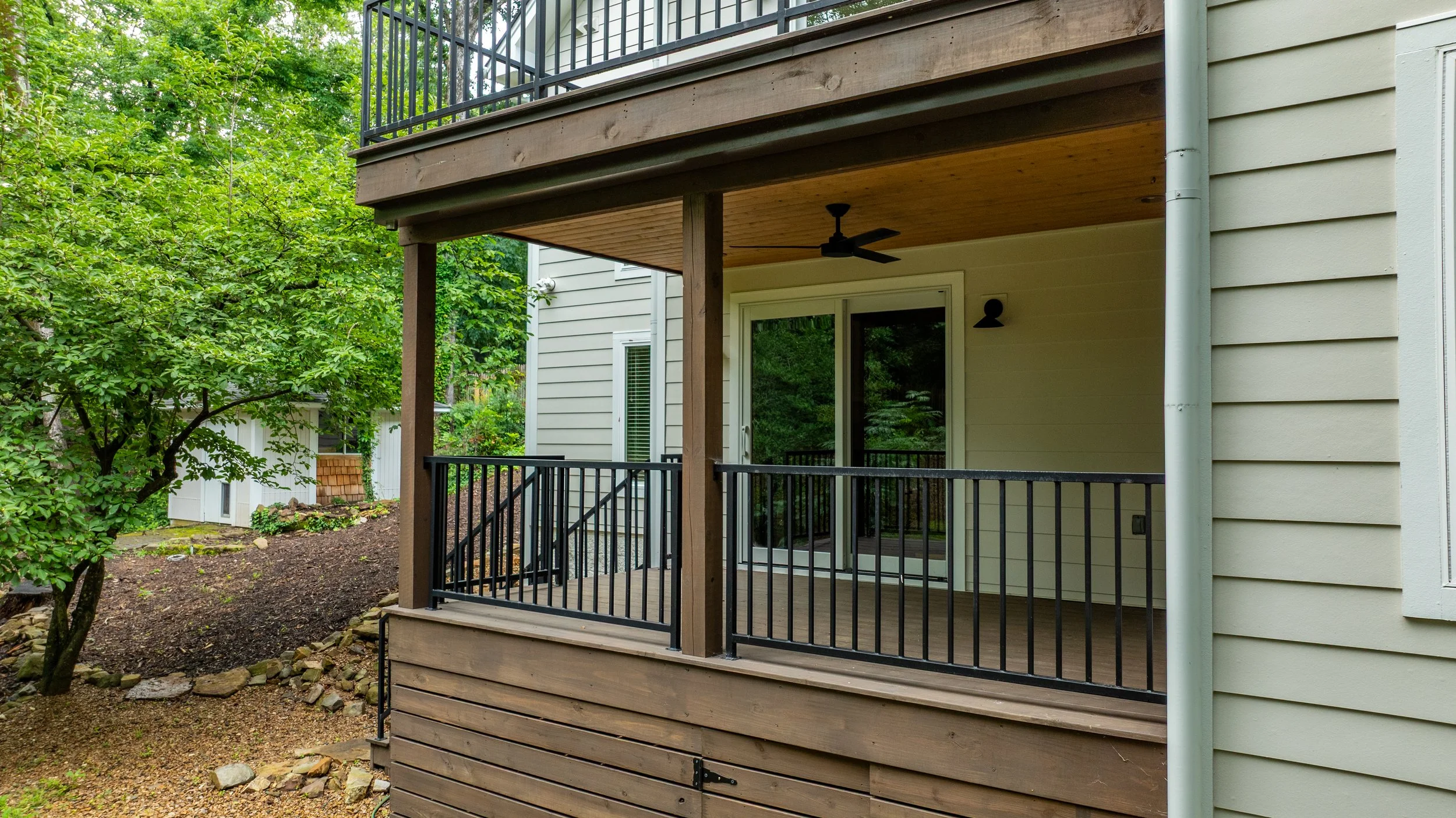 Back porch of a house with black metal railing, wooden ceiling, ceiling fan, sliding glass door, and beige siding, surrounded by green trees and landscaping.