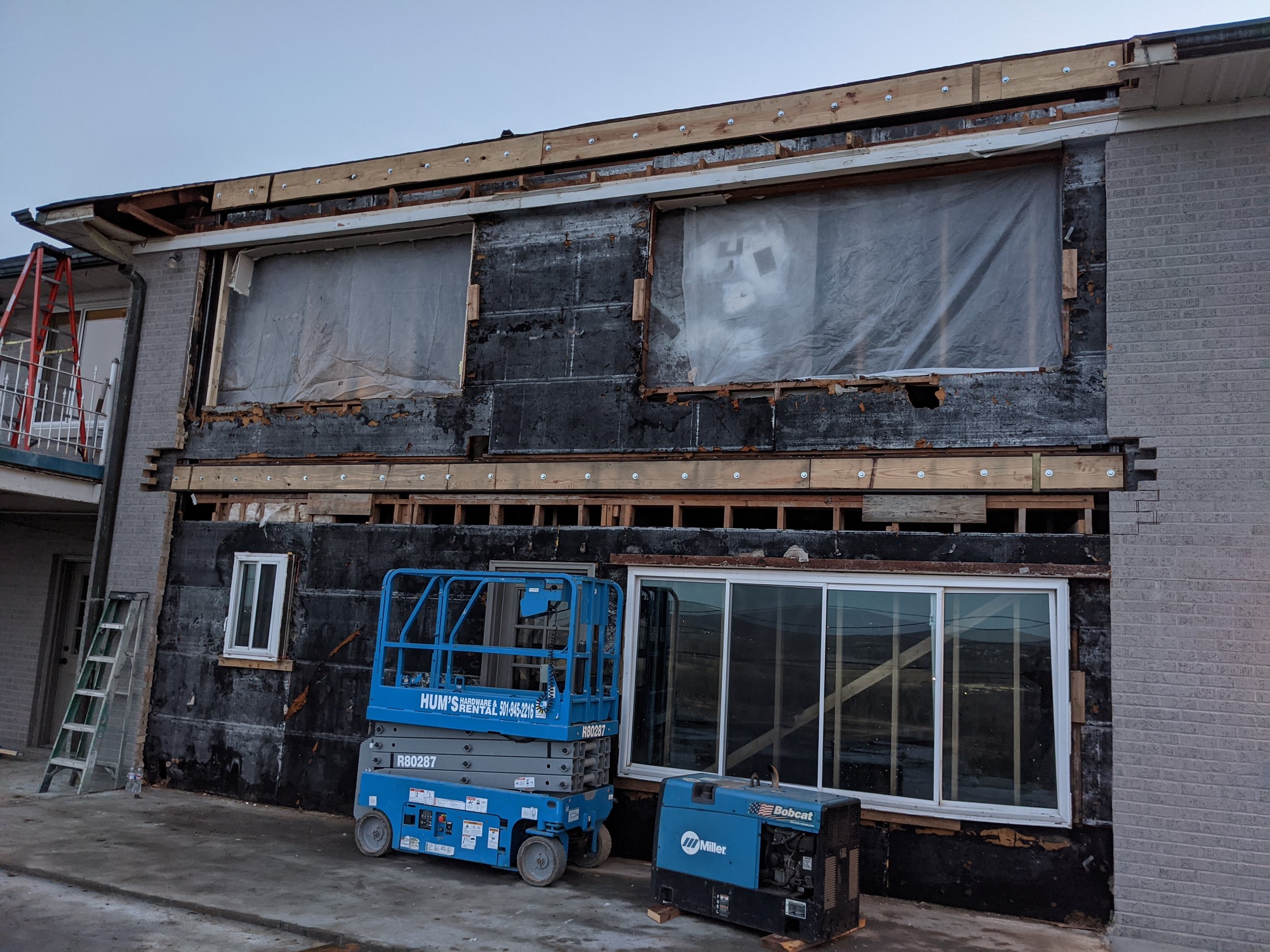 Construction site with building undergoing renovation; partially removed exterior wall, insulation, and plastic sheeting covering window openings; construction lifts, ladder, and tools in the foreground.
