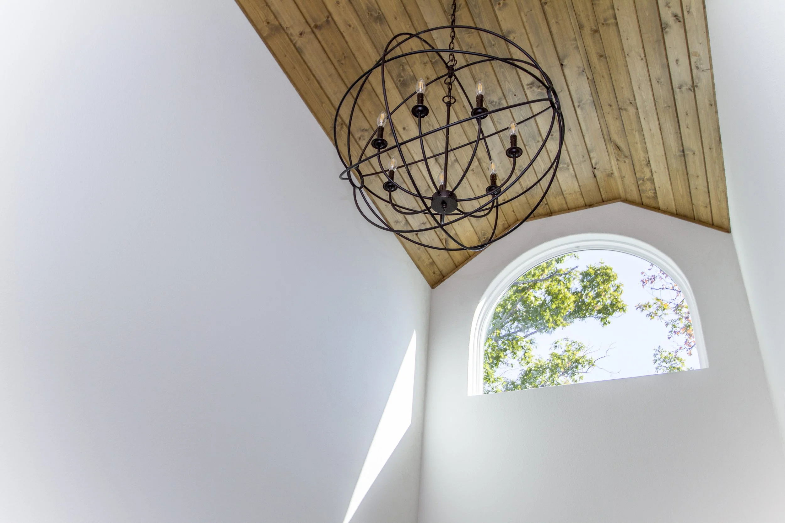 Interior view of a room showing a ceiling with wooden paneling, a modern chandelier, a large arched window with trees outside, and white walls.