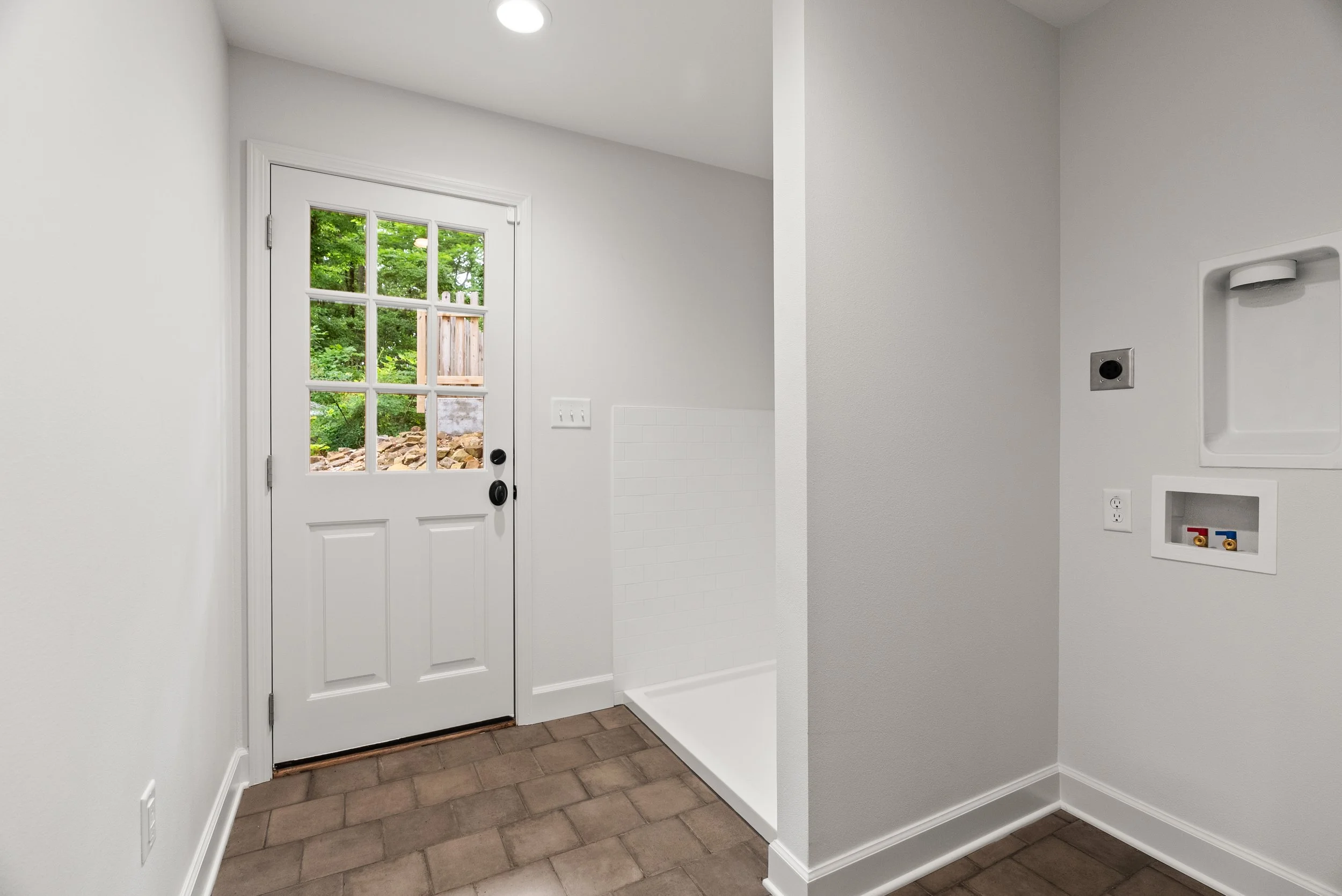 Interior of a modern laundry or utility room with white walls, a door with glass panels showing greenery outside, and connections for a washer and dryer.