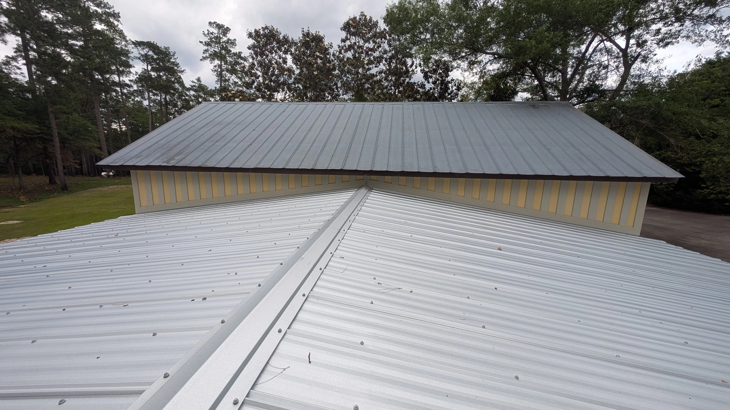 A metal roof with the camera pointing towards a ridge, showing a second metal roof with a different color in the background surrounded by trees.