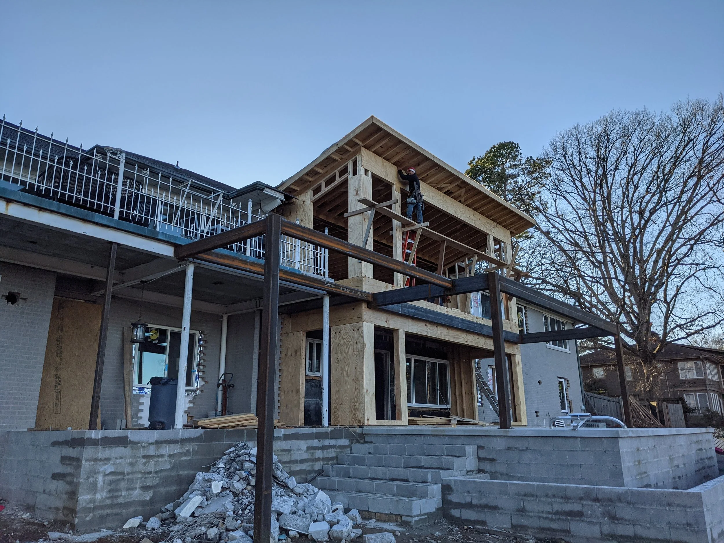 Construction workers building a second story wooden extension on a residential house, with a staircase and rubble at the ground level, in the late afternoon.