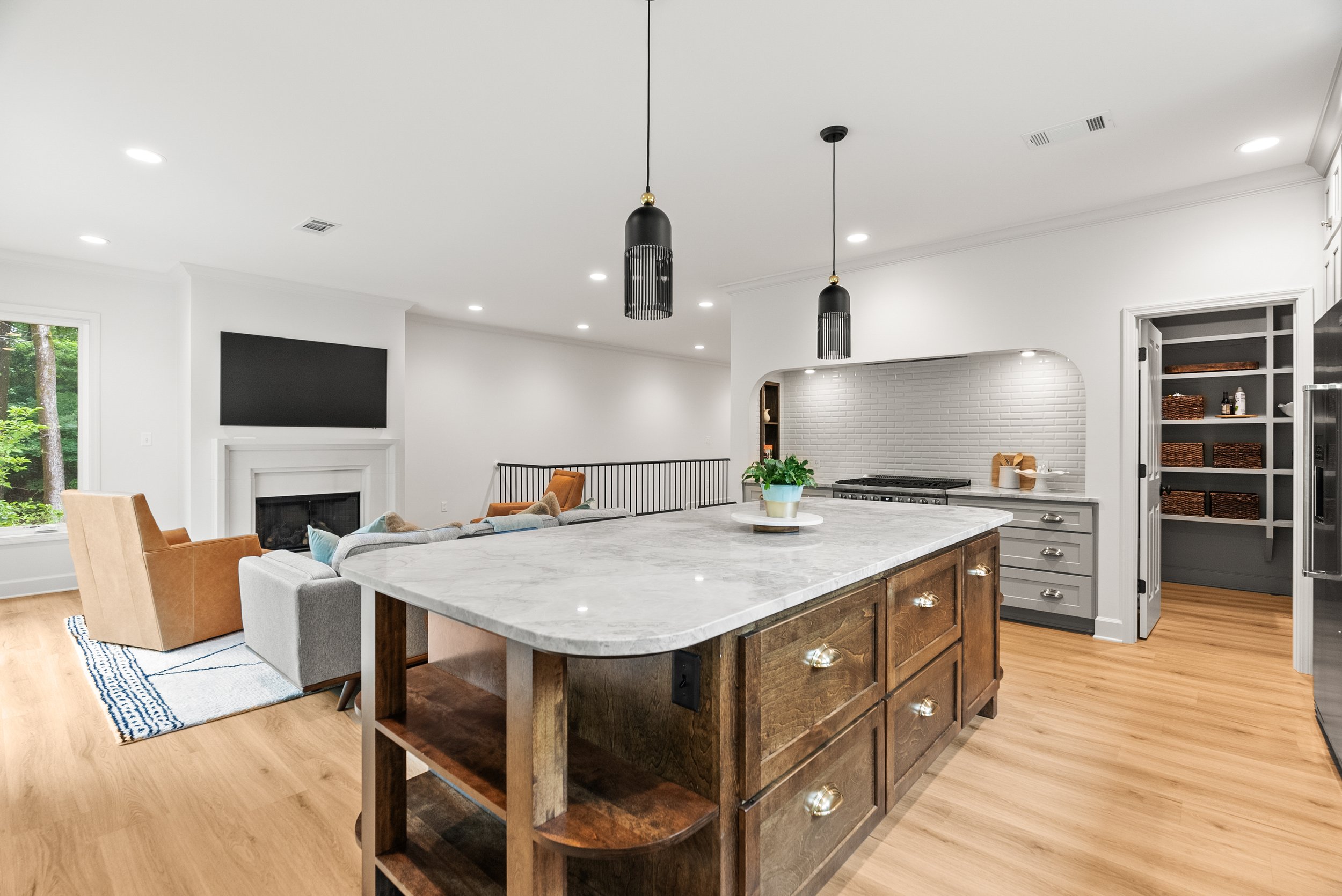 Open-concept kitchen and living room with white walls, hardwood floors, and modern decor. Kitchen features a white marble island, dark wood cabinets, and stainless steel appliances. Living room has a sectional sofa, a fireplace, and wall-mounted TV.