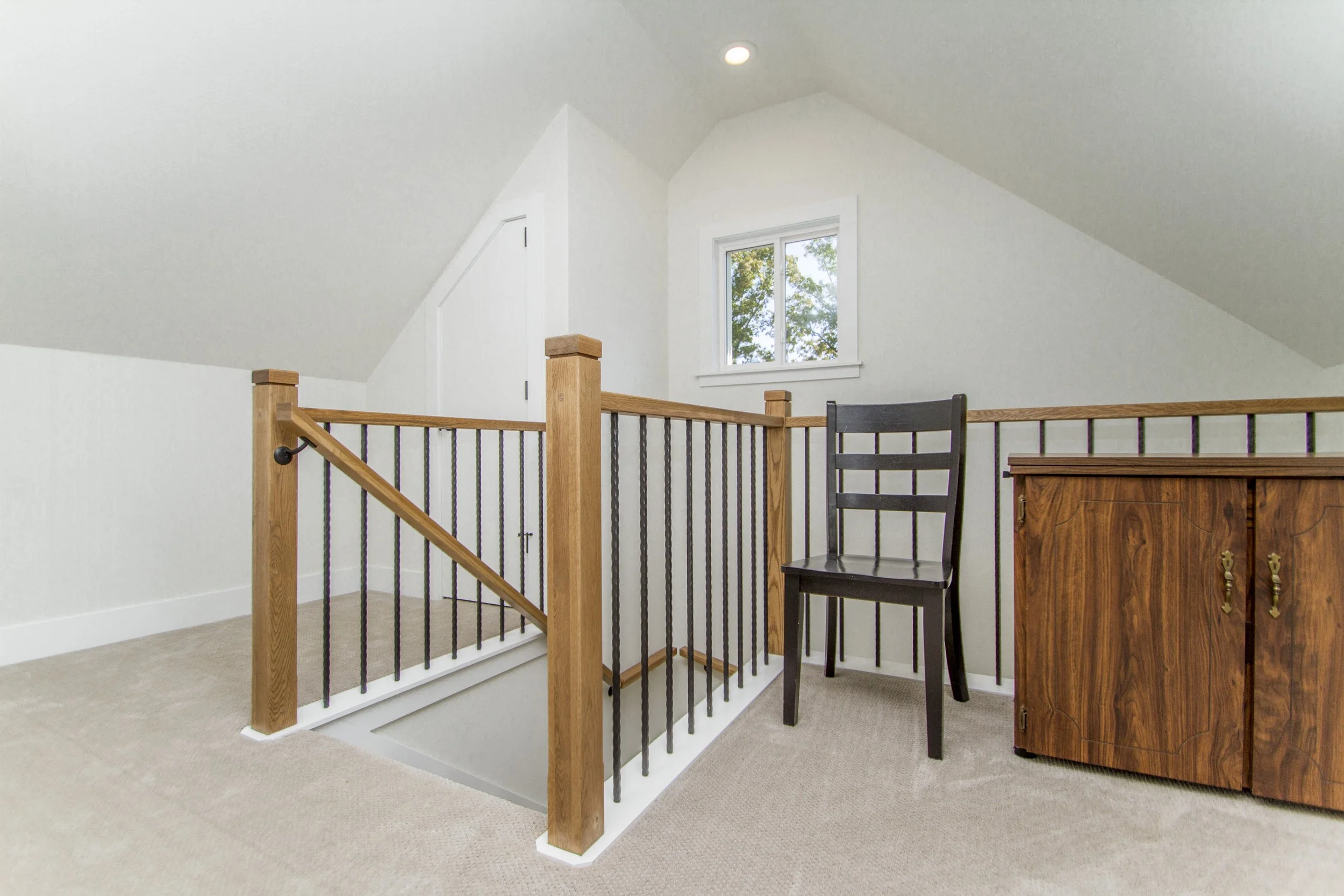 Interior of a room with a staircase that has wooden posts and black metal balusters, a black chair, a wooden cabinet, carpeted floor, and a window showing trees outside.