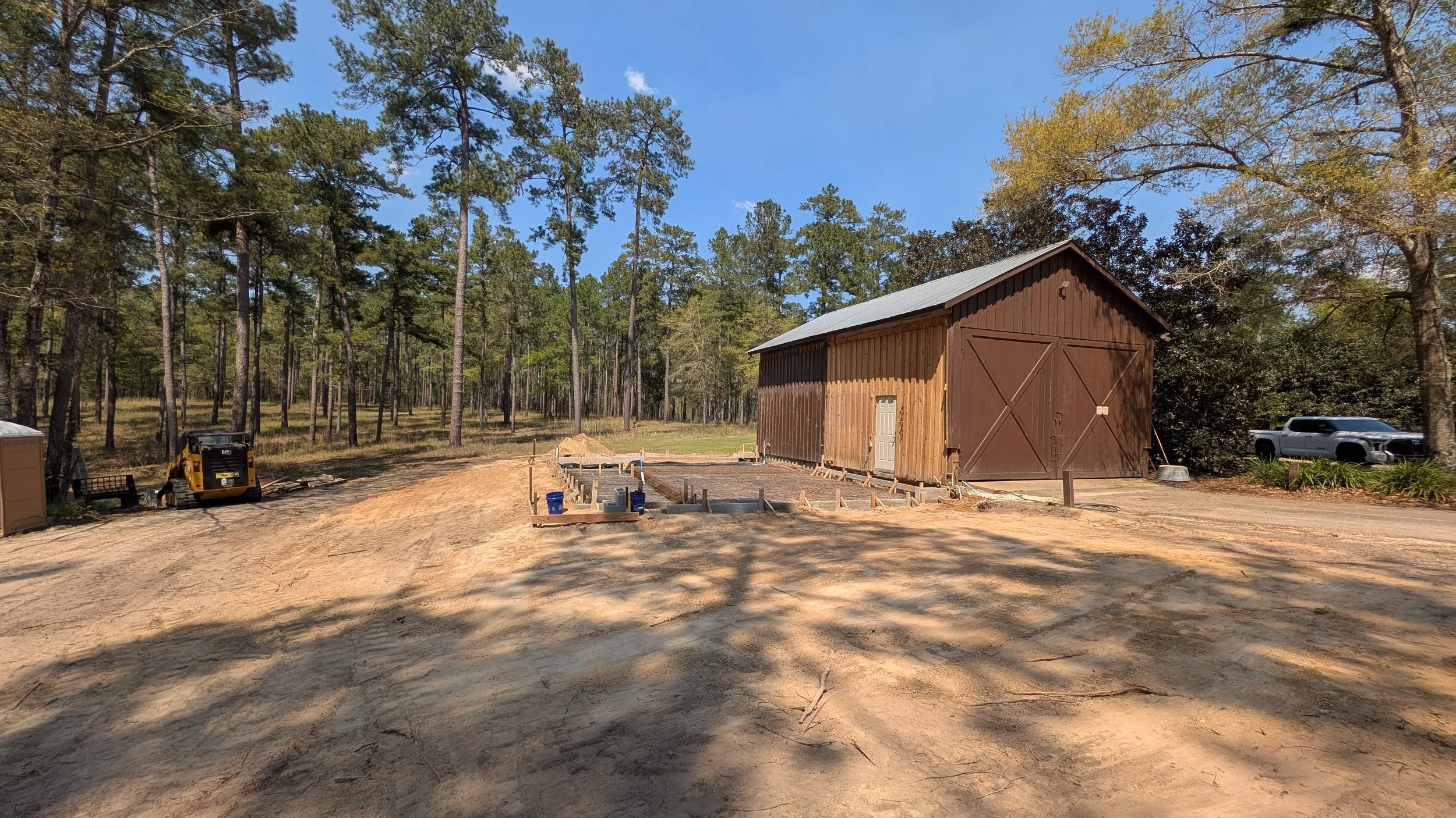Construction site with a wooden barn, foundation work in progress, and construction equipment surrounded by trees under a blue sky.
