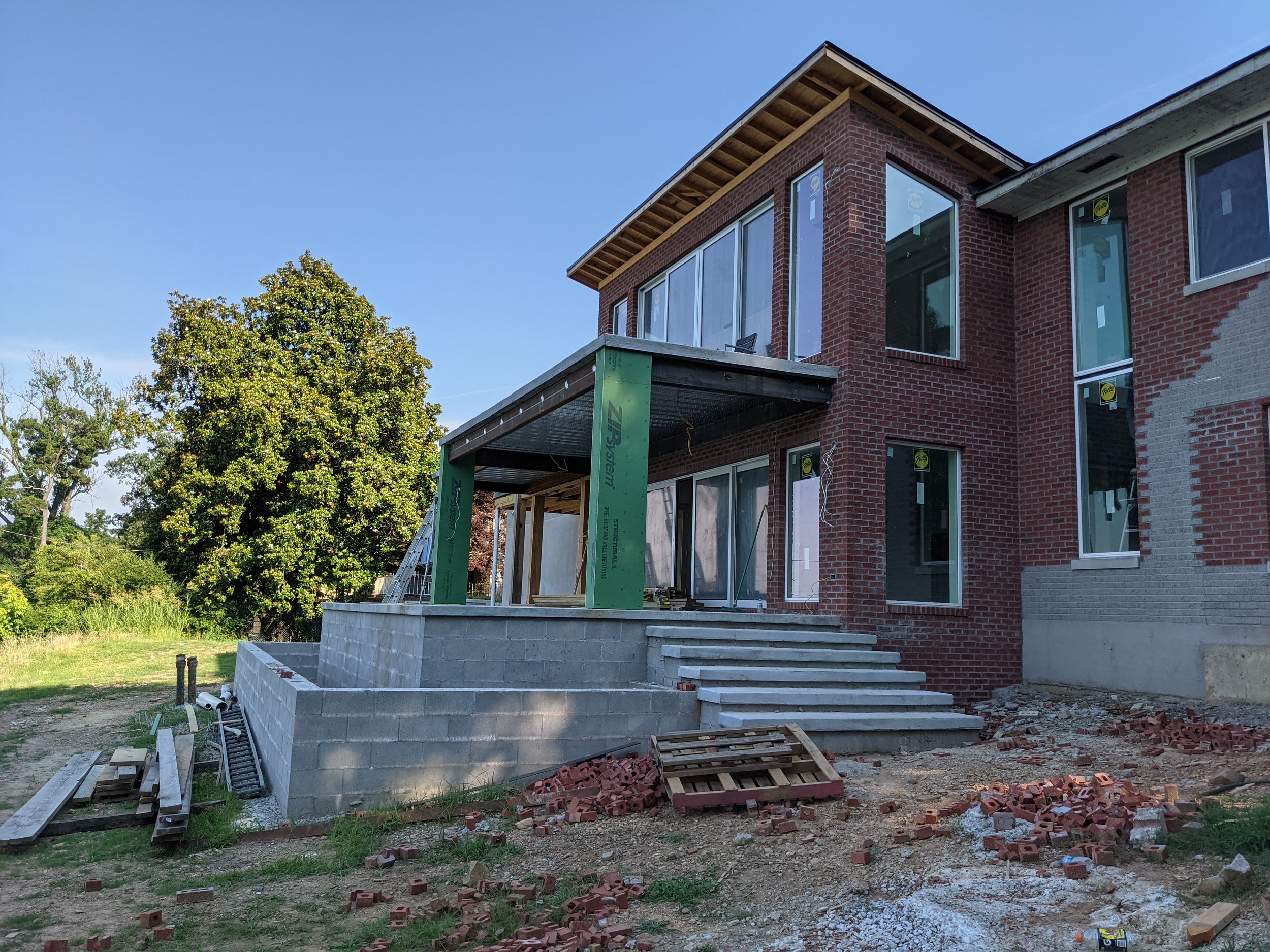 A house under construction with brick exterior, large windows, and a partially built deck with concrete blocks and construction materials on the ground.