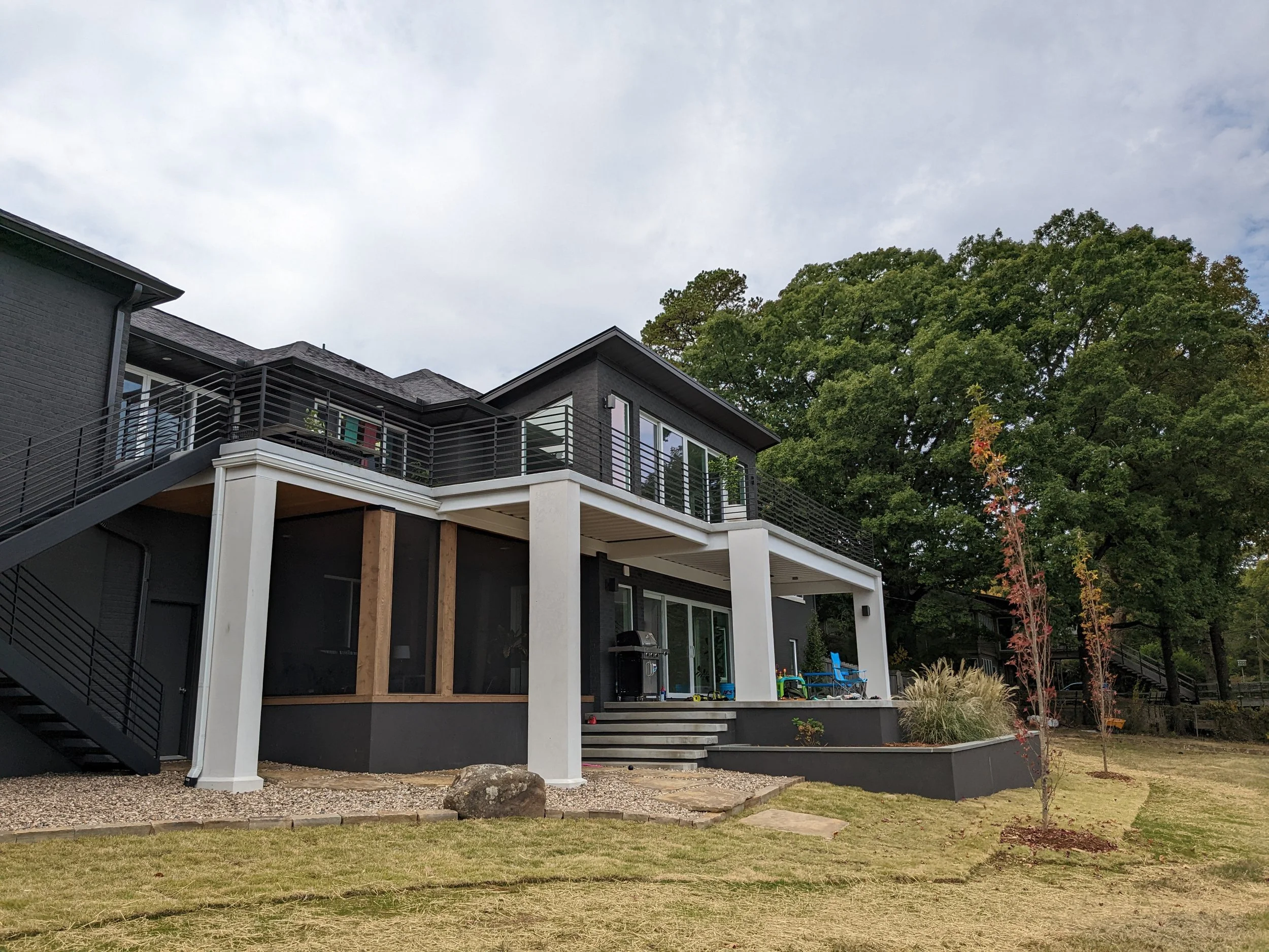 Modern multi-story house with dark gray siding, large glass windows, balcony with black railing, surrounded by yard with small trees and plants, under a cloudy sky.