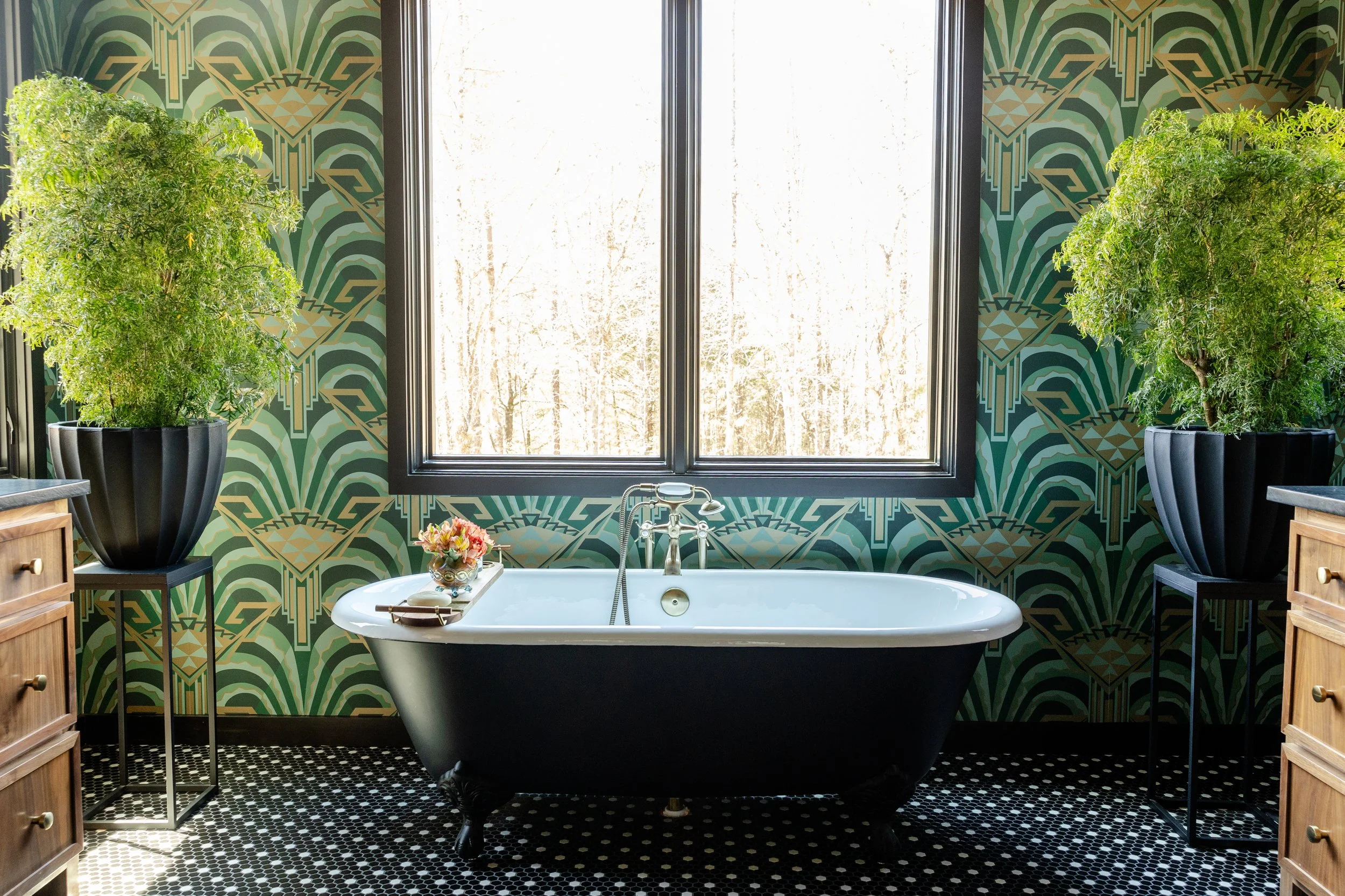 Bathroom with a vintage clawfoot bathtub in front of a window, flanked by two large potted plants. The wall has green geometric patterned wallpaper, and wooden cabinets are on either side of the tub. There's a small table with flowers and bathing ess