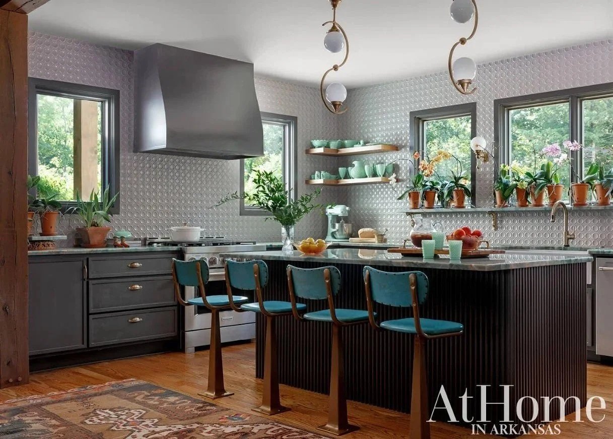 A modern kitchen with gray cabinets, pegboard backsplash, and wooden floors. It features a kitchen island with four blue chairs, potted plants on windowsills, and open shelves with teal dishes. The room has three windows letting in natural light and a hanging light fixture with globe bulbs.