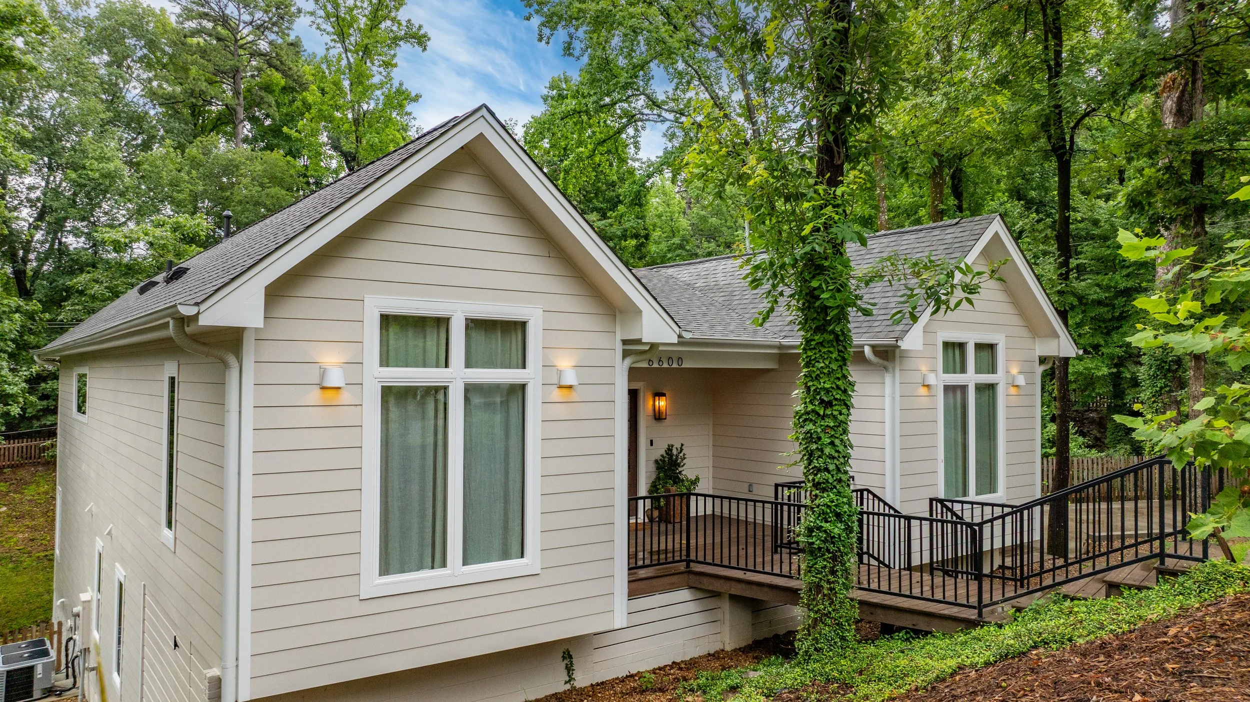 A modern two-story house with beige siding and white trim, surrounded by green trees, featuring large windows, a small porch with black railing, and outdoor lighting.