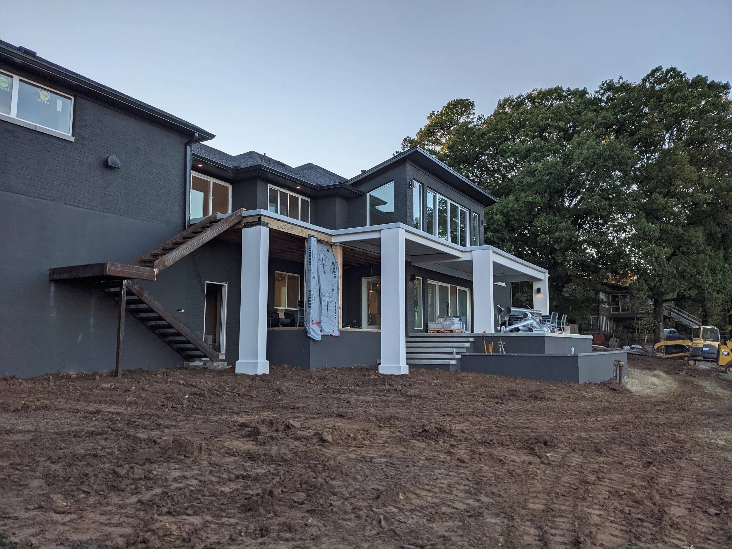 Rear view of a two-story house under construction with a modern design, black exterior walls, large glass windows, a balcony, and a construction area with earth and construction equipment in the foreground.