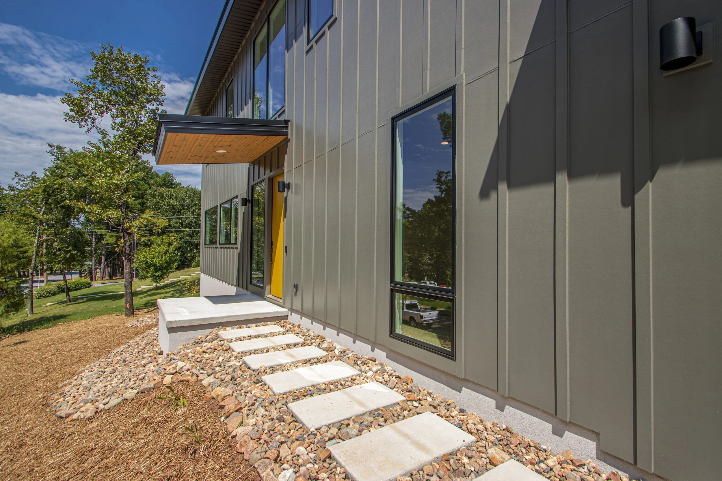 Exterior of a modern two-story building with large windows, a yellow front door, a small concrete porch, and a pathway with stepping stones leading to the entrance. The building has vertical metal siding and is surrounded by landscaped ground with mu