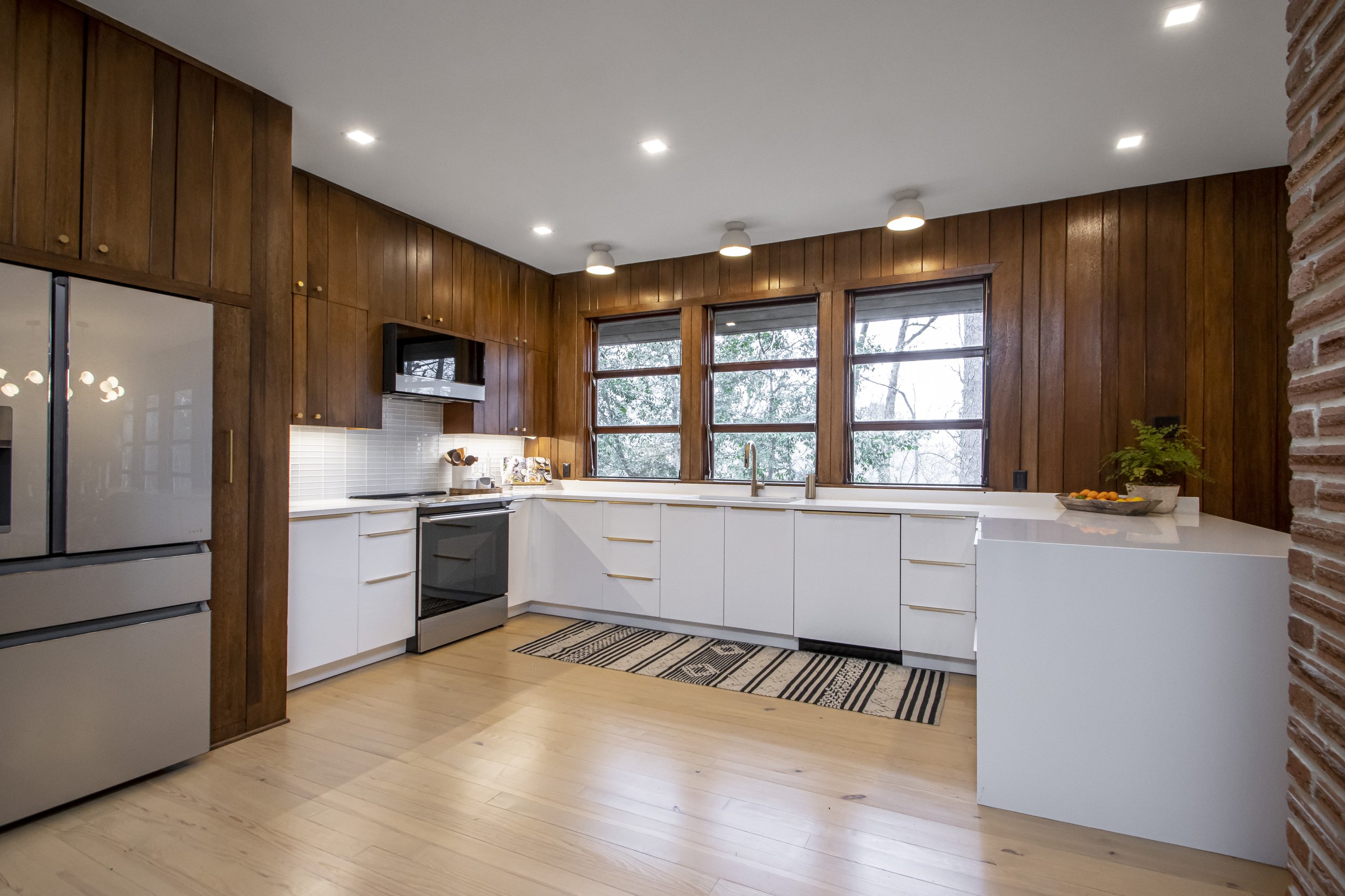Modern kitchen with white cabinets, black appliances, and wooden wall paneling, large windows, and recessed ceiling lights.