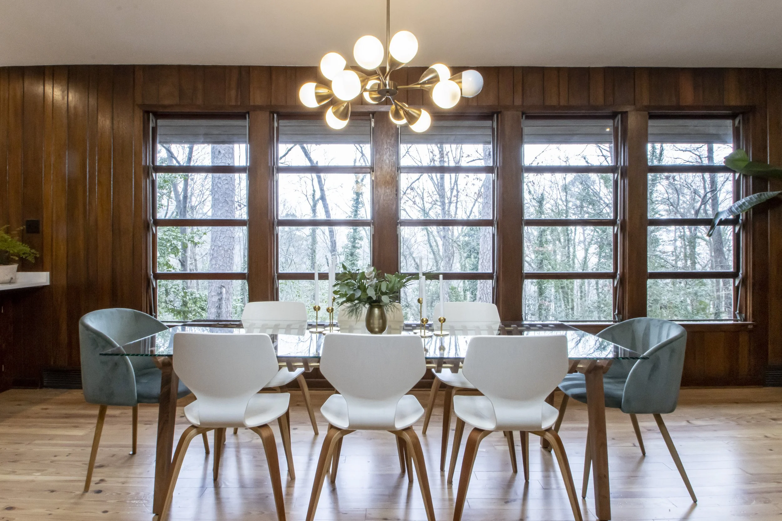 A dining room with wooden walls and large windows showing a view of trees outside. There is a rectangular glass table surrounded by white and dark blue chairs. On the table, there is a green plant in a gold vase and gold candlesticks. An overhead lig