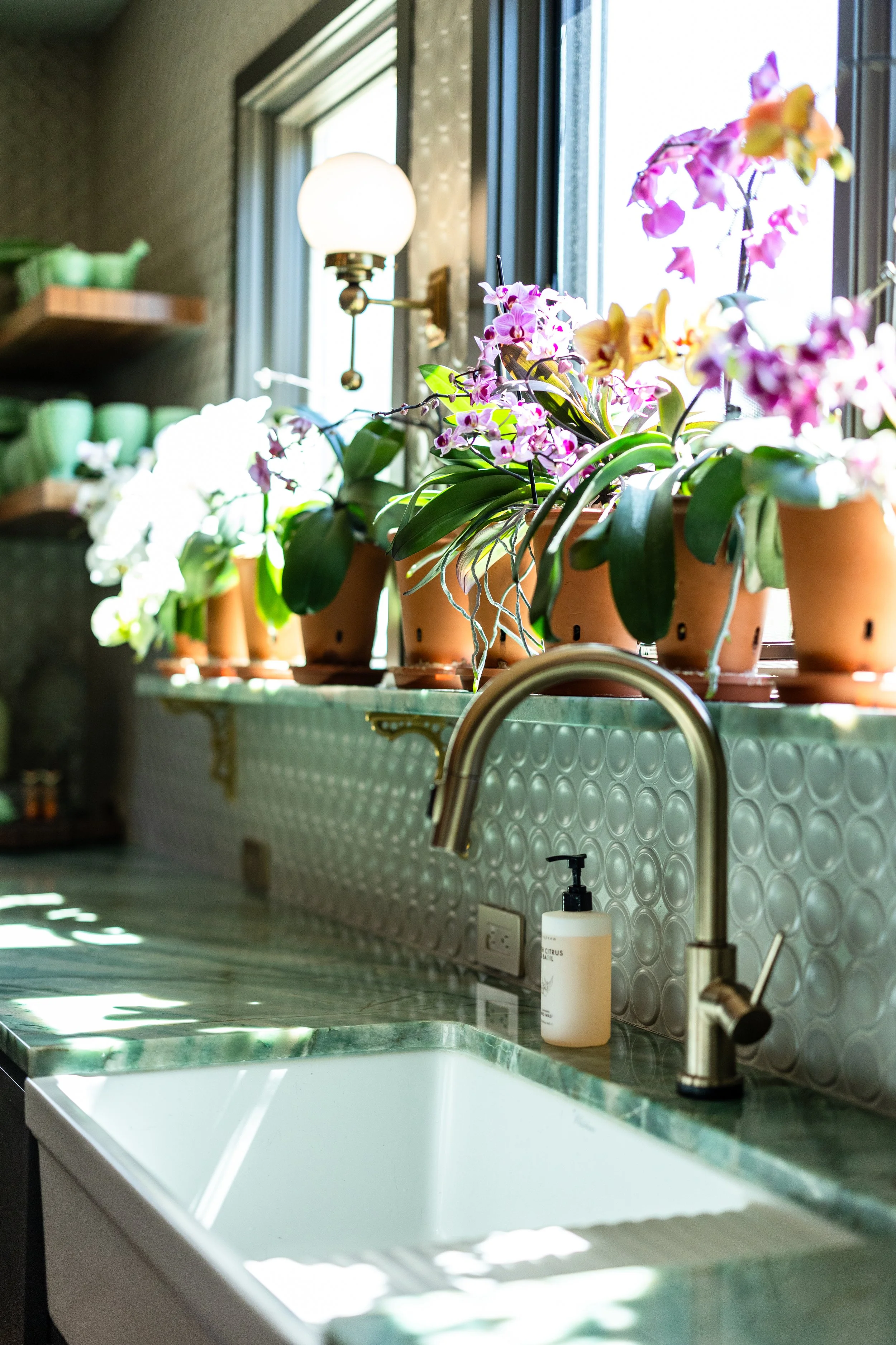 Potted orchids on a green marble kitchen countertop near a white sink, with a window and a wall-mounted light fixture in the background.