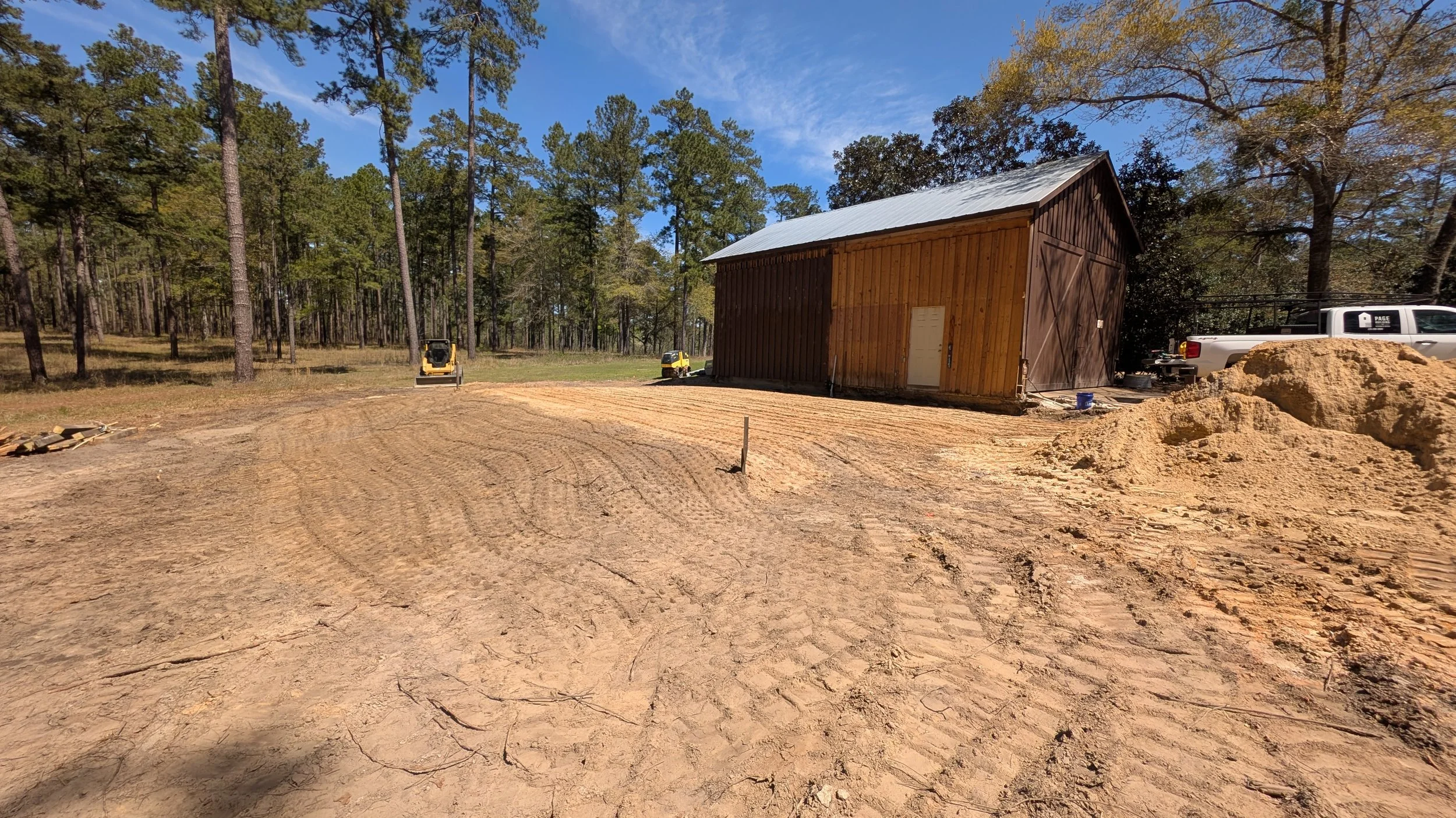 A wooden barn under construction in a cleared area surrounded by tall pine trees, with construction equipment and a pickup truck nearby, and a dirt ground with visible tire tracks.