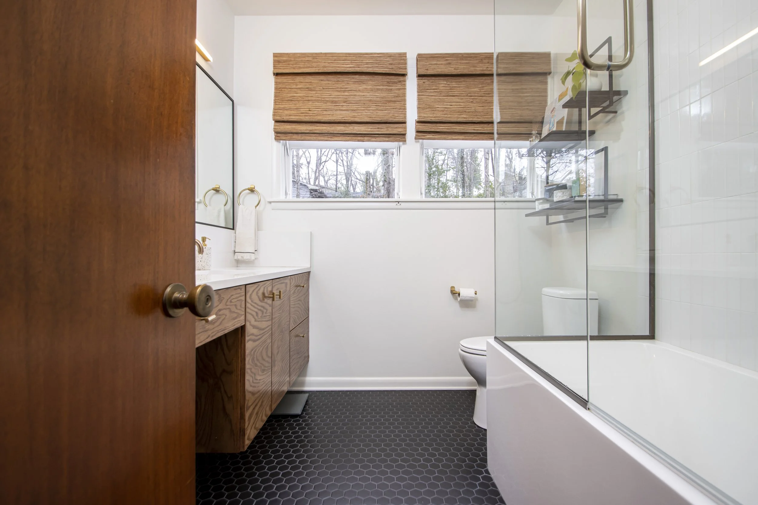 A modern bathroom with a wooden vanity, a rectangular mirror, a white toilet, a tiled shower with glass door, and windows with brown blinds showing trees outside.