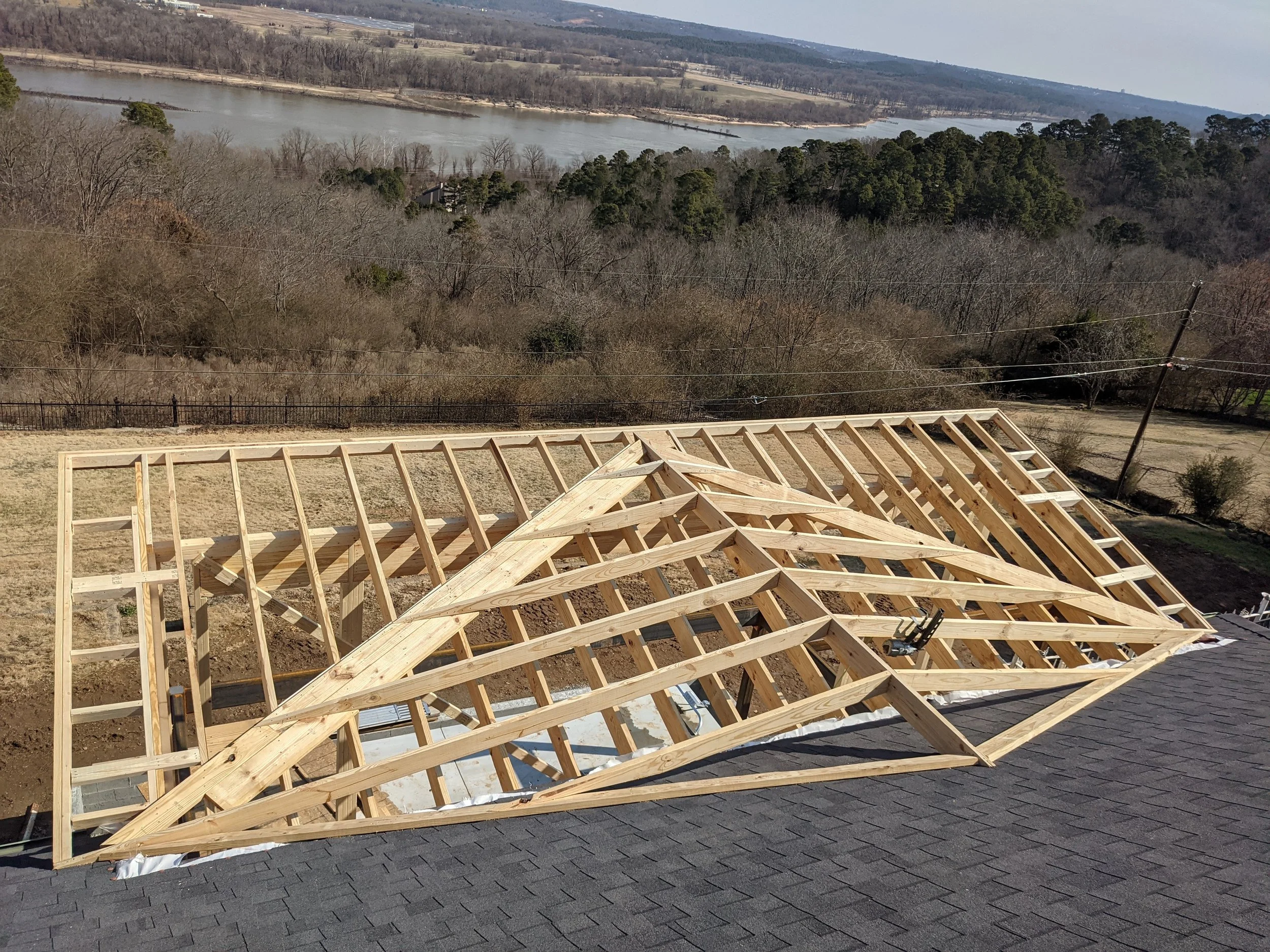 Wooden roof framing structure under construction with a scenic view of a river, hills, and trees in the background.