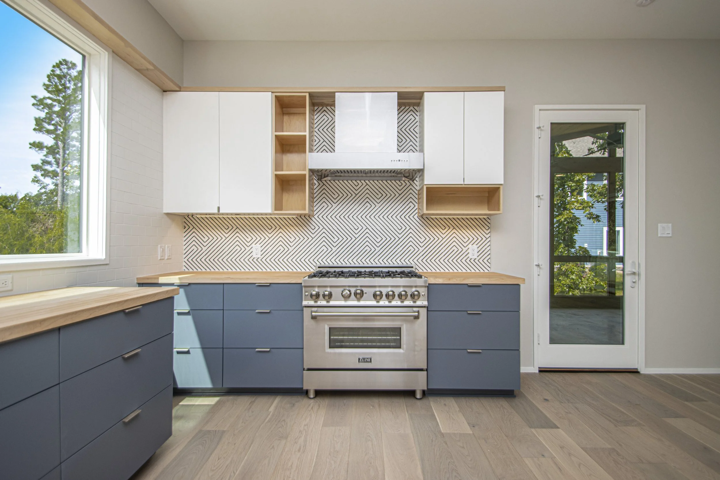Modern kitchen with blue lower cabinets, wooden countertops, a stainless steel oven, and a decorative black-and-white geometric backsplash. Two large windows and a glass door provide natural light.