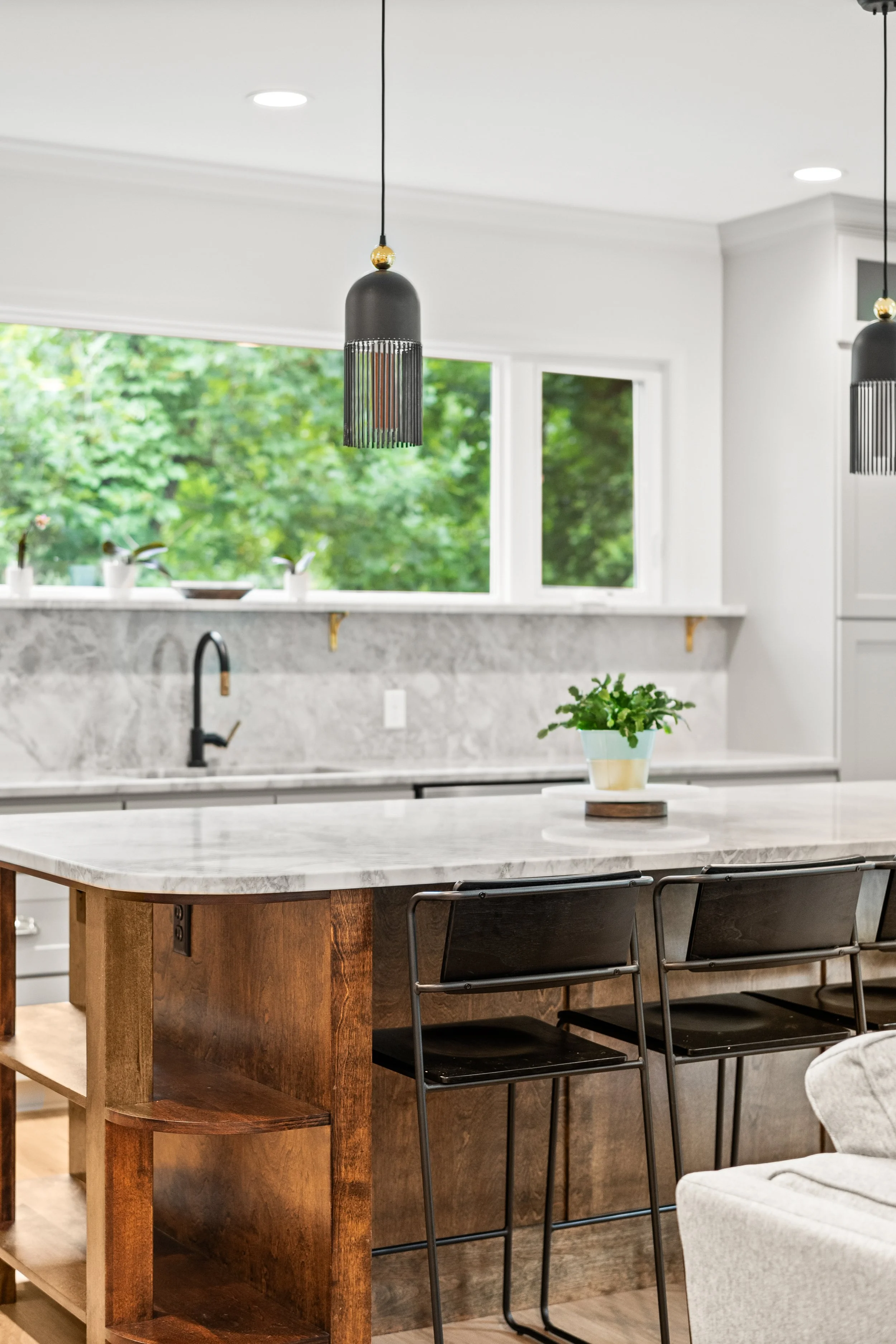 Inside a modern kitchen with a large white marble island, black modern pendant lights, green plants in small pots near the window, and black chairs around the island.