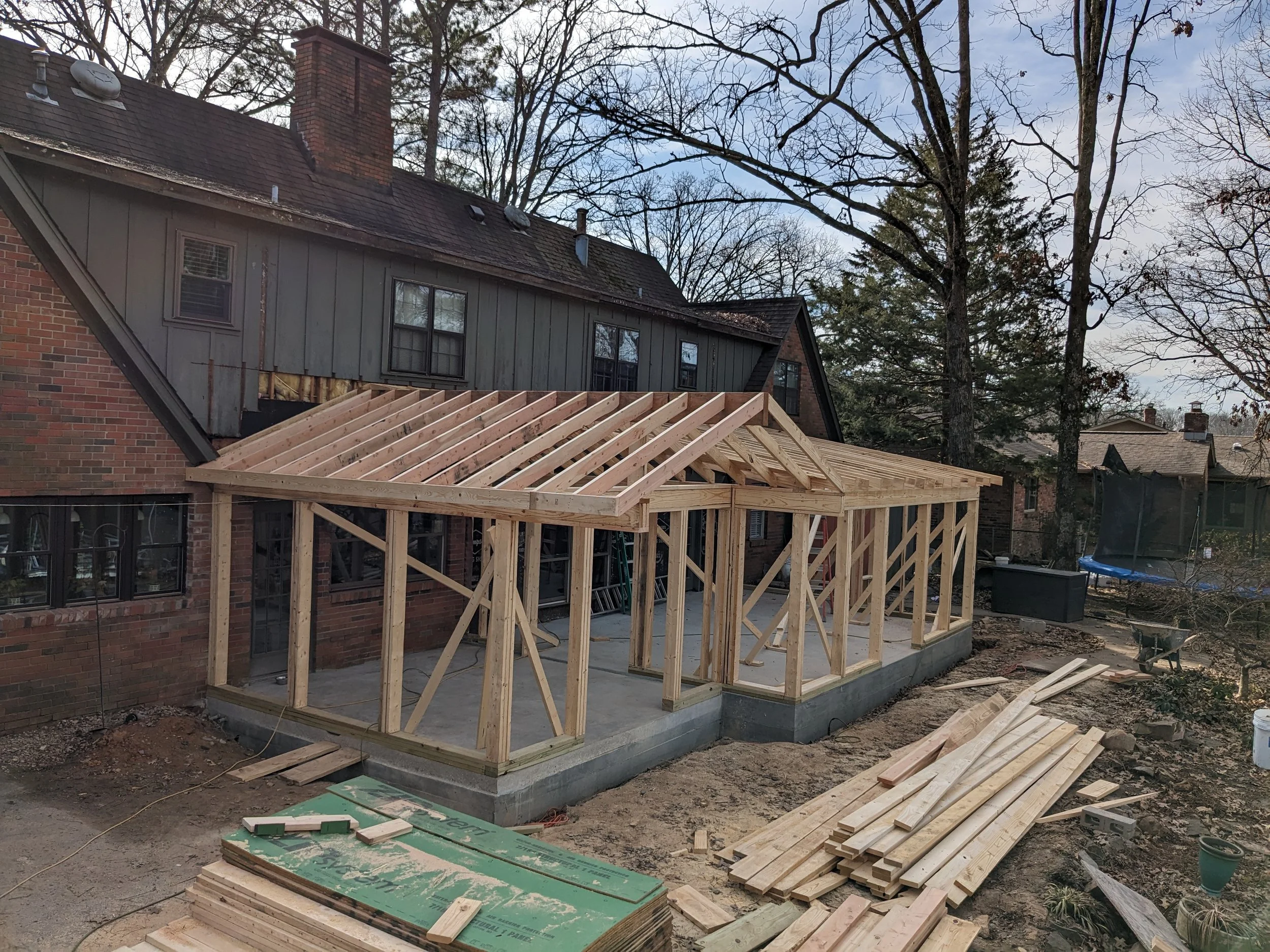 Construction site of a backyard addition with a wooden frame foundation and walls on a concrete slab in front of an existing brick and wood house, with trees and roof visible in the background.