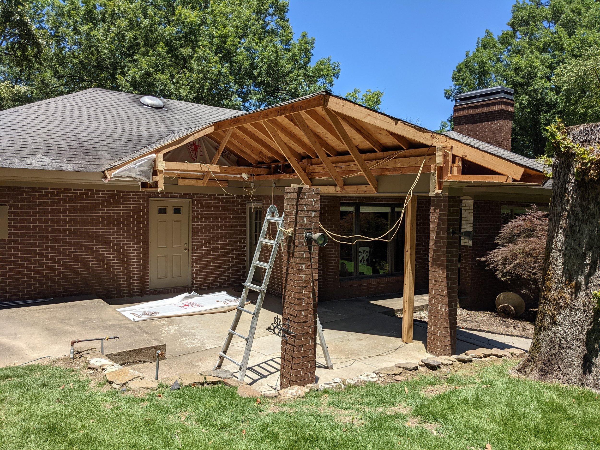 A house undergoing roof extension with wooden framing, a ladder, construction tools, and scaffolding, surrounded by trees and a clear blue sky.