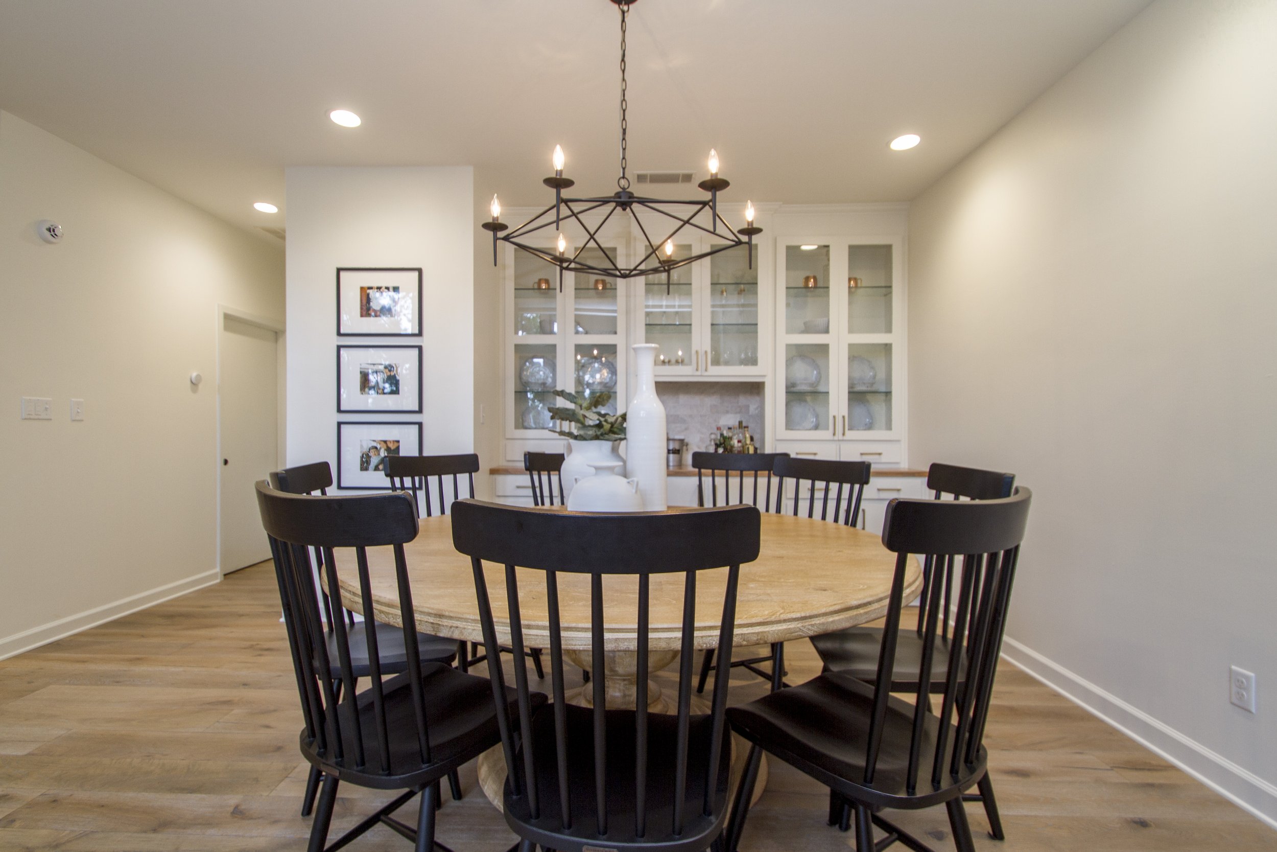 A dining room with a round wooden table surrounded by black wooden chairs. The room features a modern black metal chandelier with candle-shaped lights hanging above the table. In the background, there is a white cabinet with glass doors displaying de