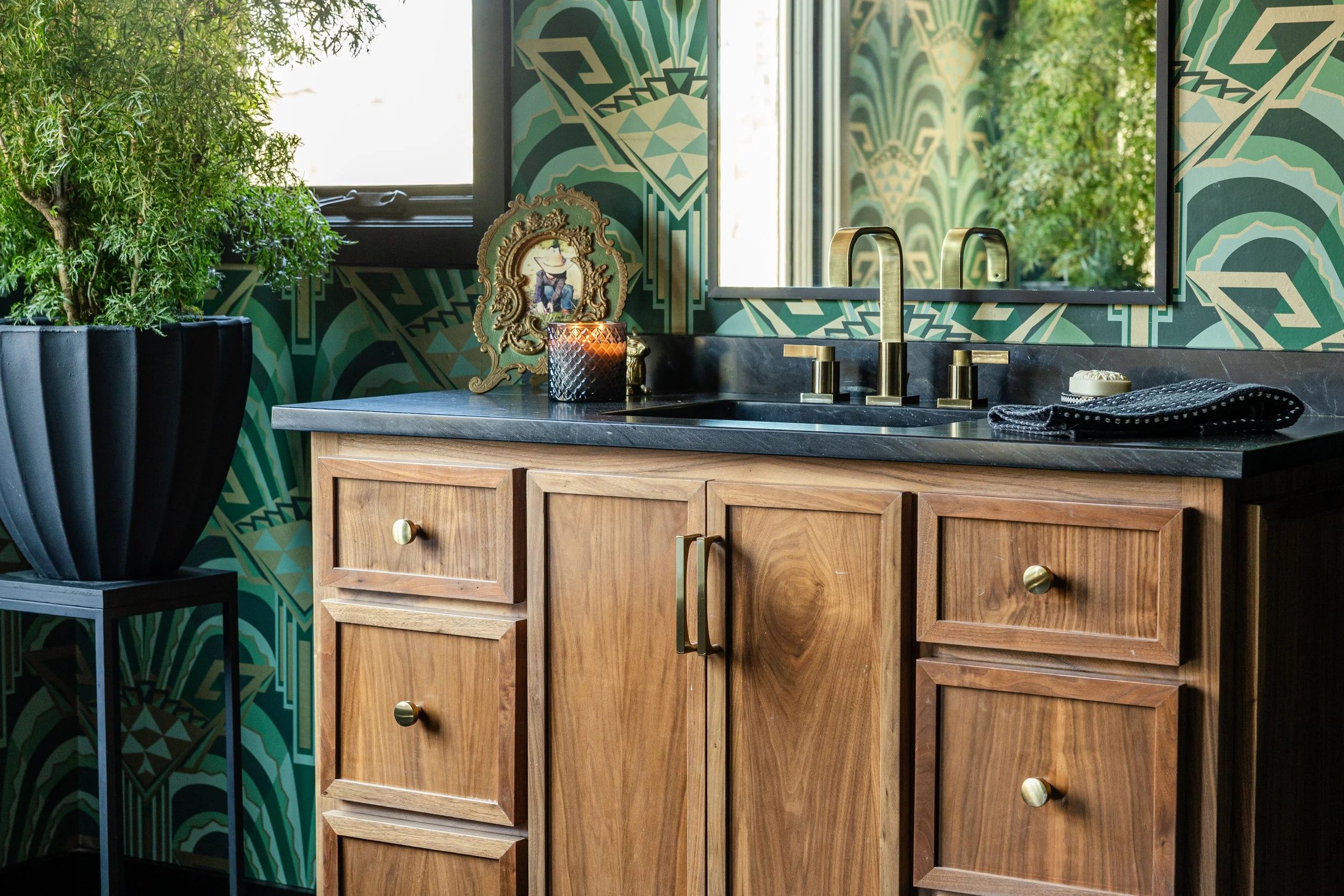 A bathroom vanity with a black countertop, brass fixtures, a decorative mirror, a candle, a framed picture, and a black towel, with green patterned wallpaper and a large window showing greenery outside.