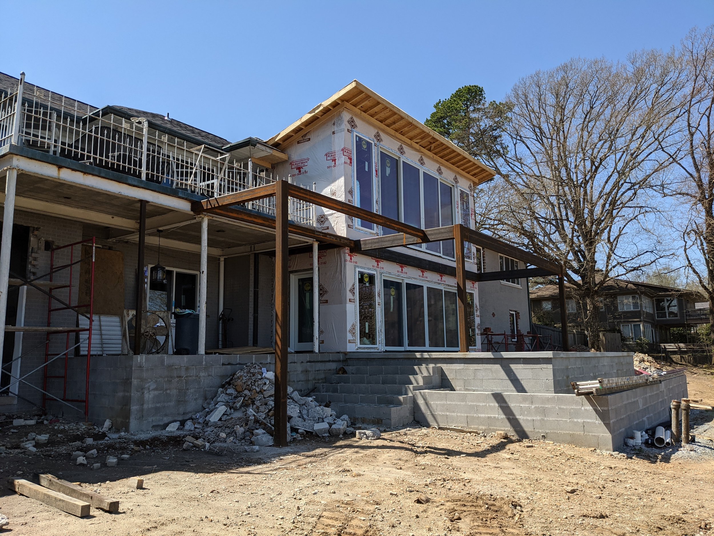 Construction site with a house under renovation, showing scaffolding, steps, and framing, with trees and a clear blue sky in the background.