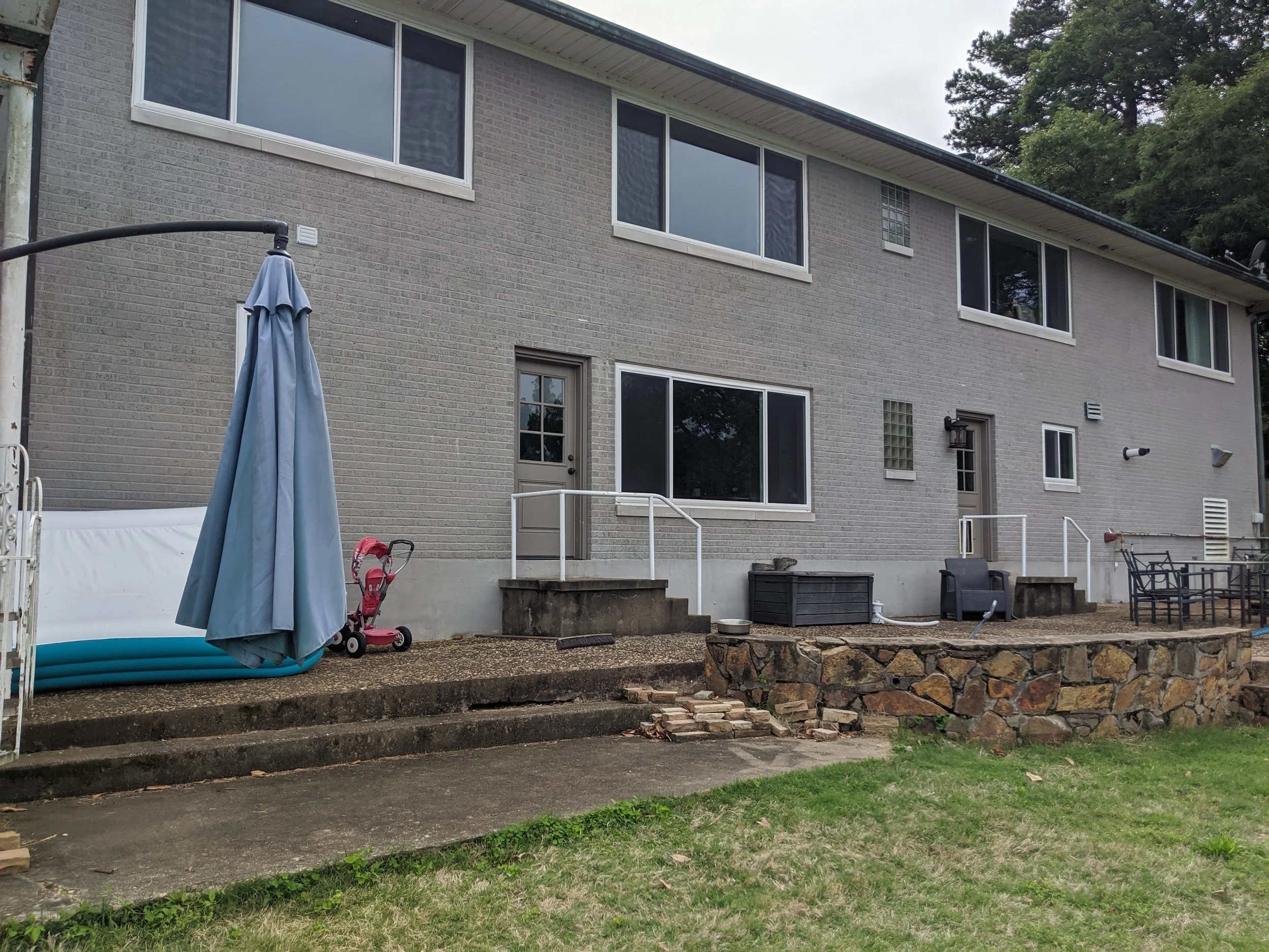 Backyard view of a gray brick two-story house with multiple windows, a patio with stairs, a blue patio umbrella, children's toys, outdoor furniture, and greenery surrounding the area.