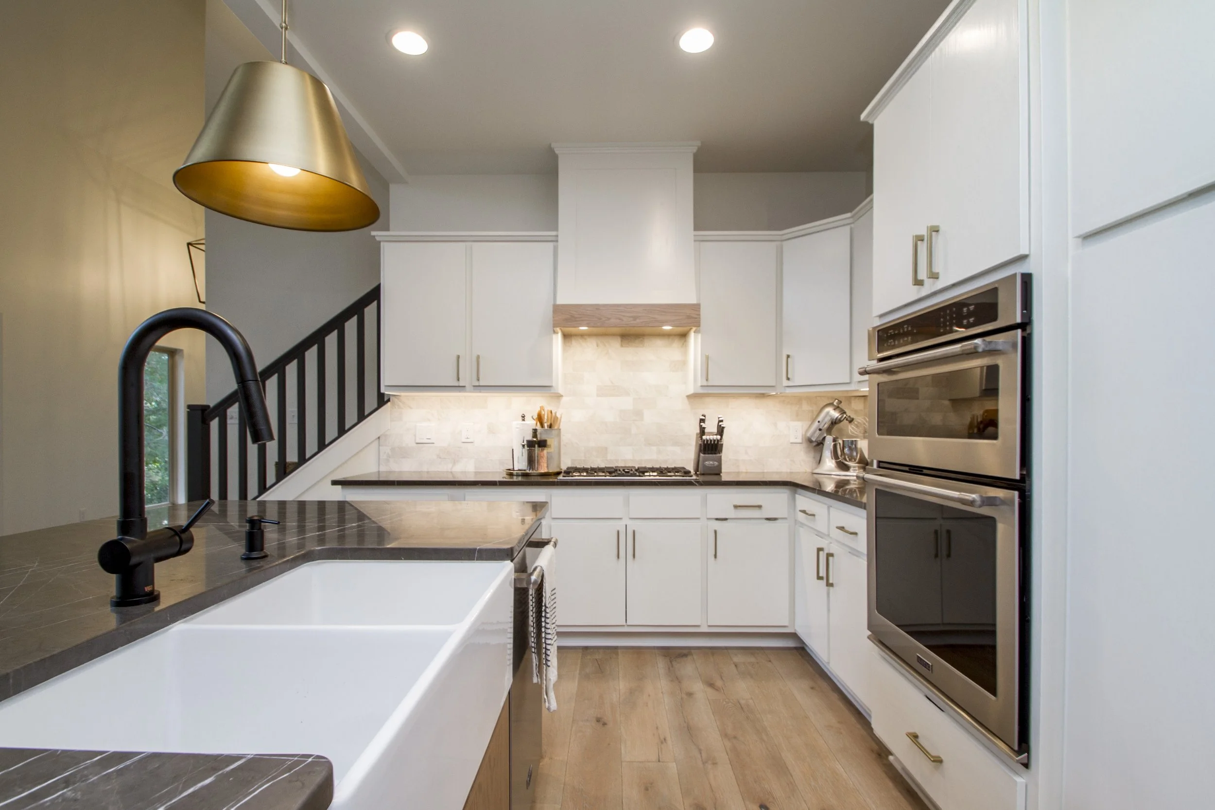 Modern kitchen with white cabinets, stainless steel oven and microwave, black countertop, beige tile backsplash, black sink faucet, and wooden flooring. Part of a kitchen island with a black marble surface and a gold pendant light overhead.