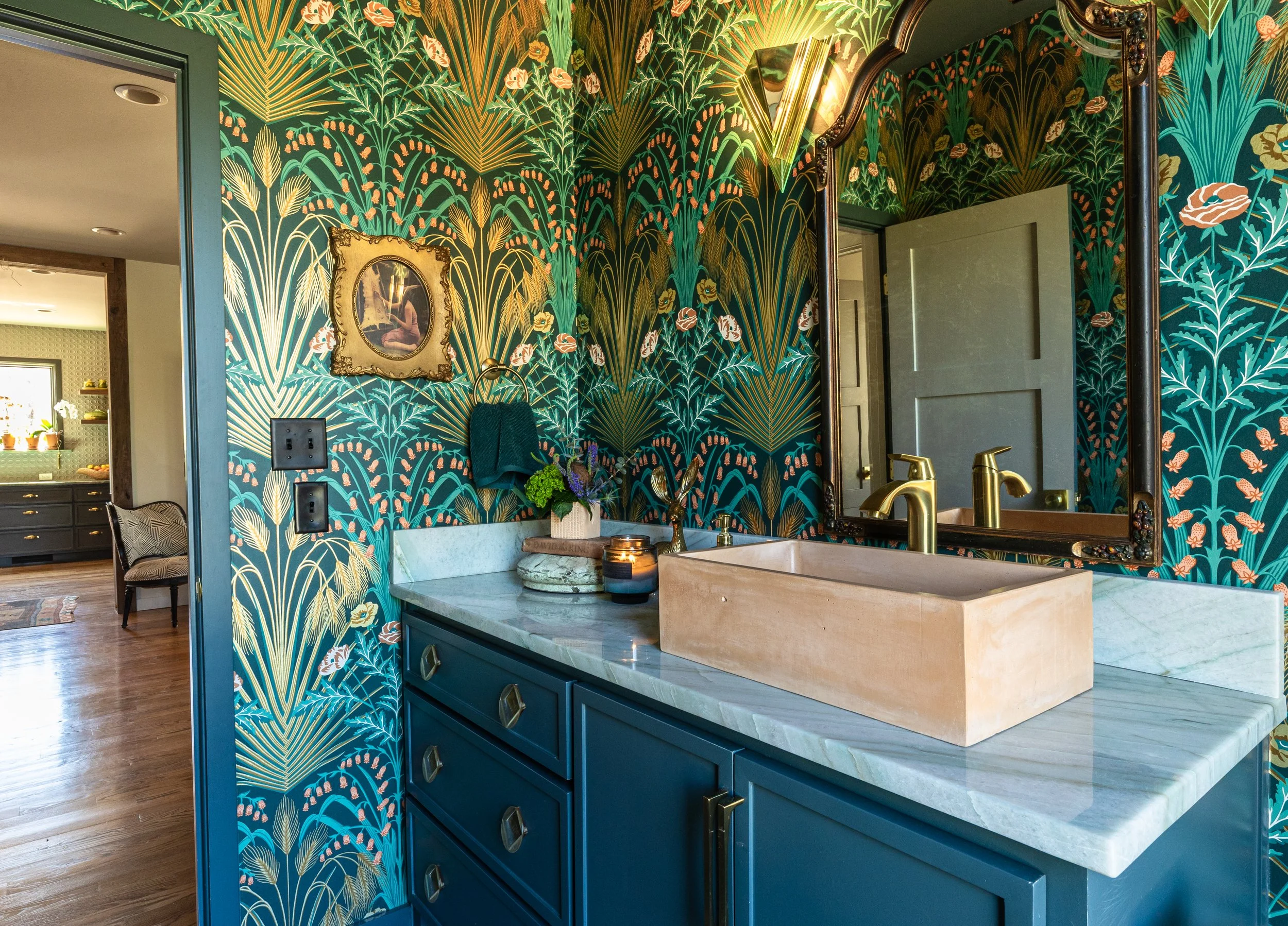 A bathroom with a floral patterned wallpaper featuring green, gold, and peach colors. A mirror with a decorative frame hangs on the wall, next to a sink with a beige stone countertop, a gold faucet, and various decorative items. To the left, part of 
