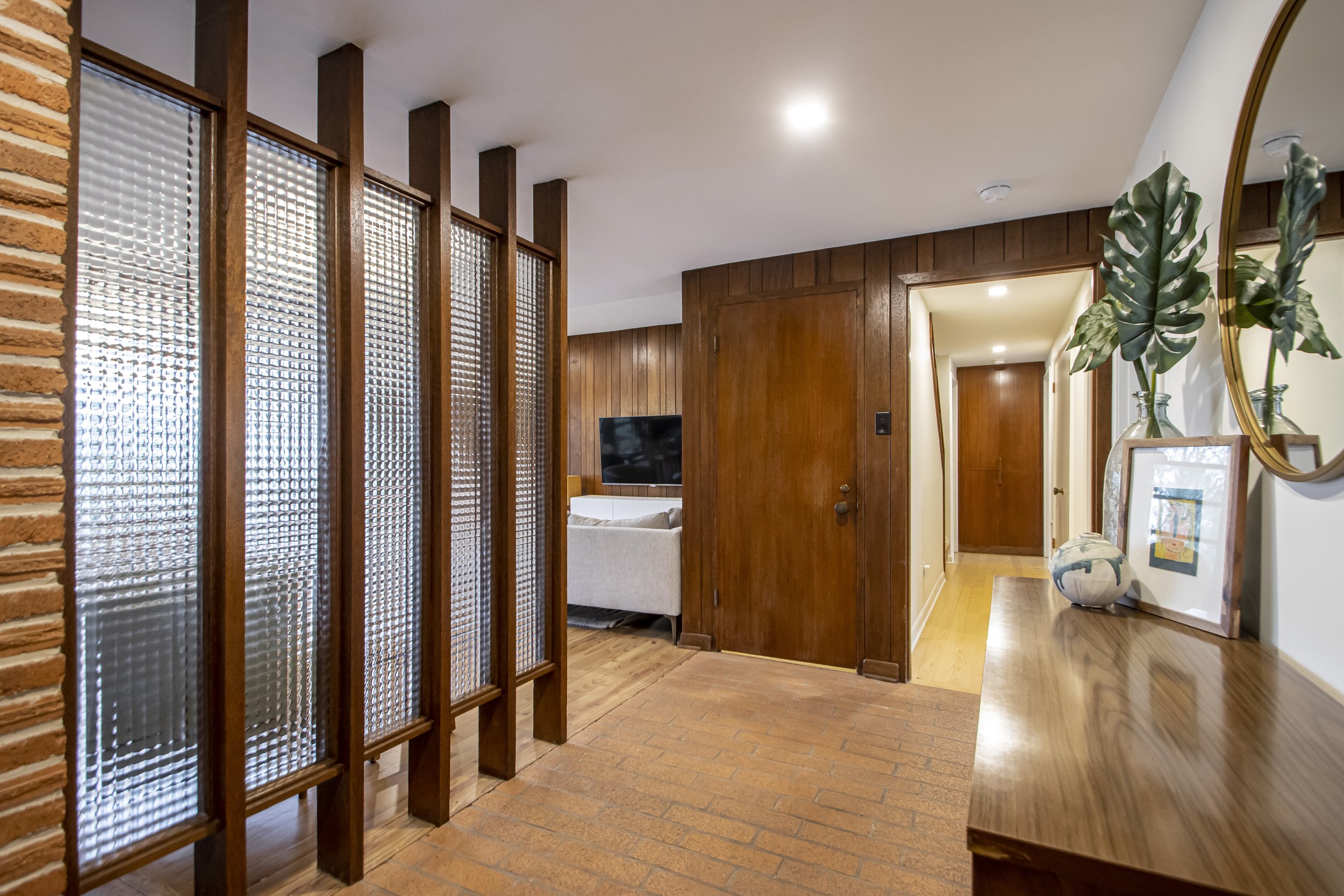Interior view of a home hallway with wooden and brick walls, a white couch, a TV, a potted plant, framed artwork, and a mirror.