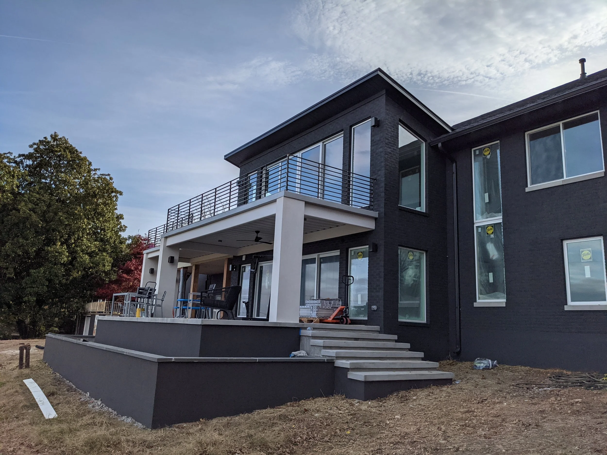 A modern house under construction with black brick exterior, large windows, a balcony with metal railings, and a covered patio area with stairs leading to the yard. There are trees and construction materials visible around the house.