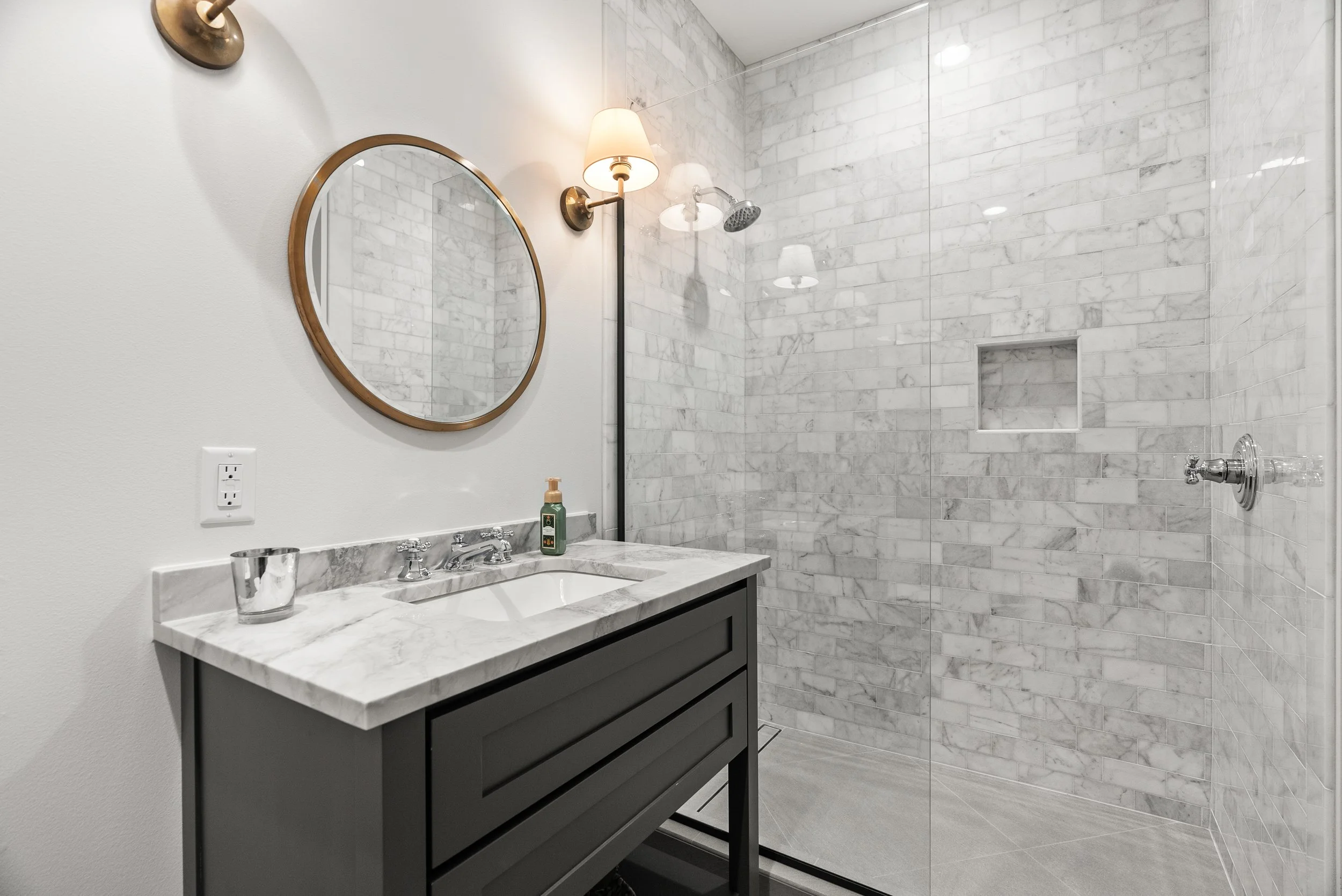 Modern bathroom with a white marble vanity, a round mirror with a gold frame, and a stall shower with marble tiles and a glass door.
