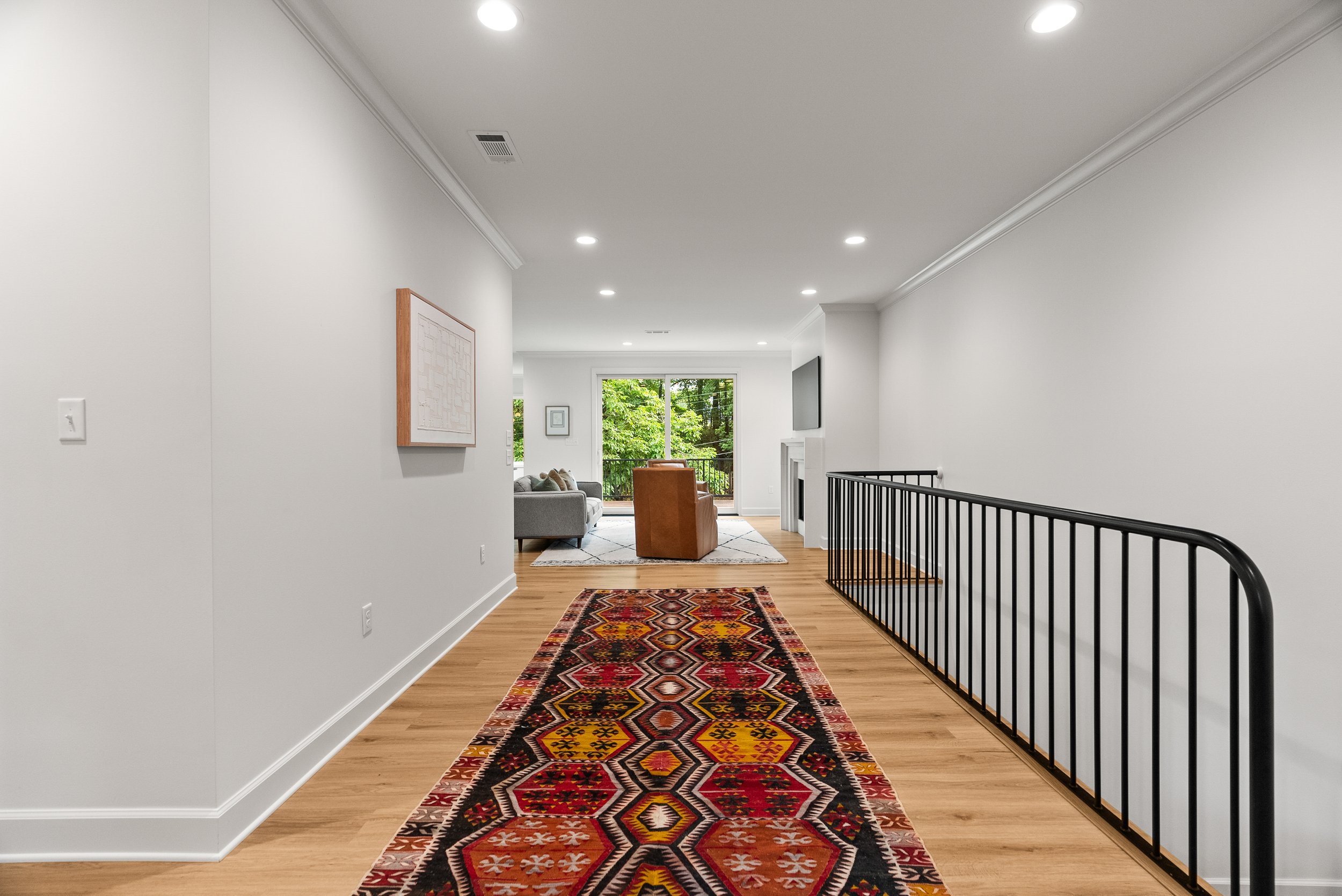 A hallway with hardwood floors, white walls, and a colorful patterned runner rug. The hallway leads to a living area with a sofa, chairs, a large window with an outside view, and a ceiling with recessed lighting.