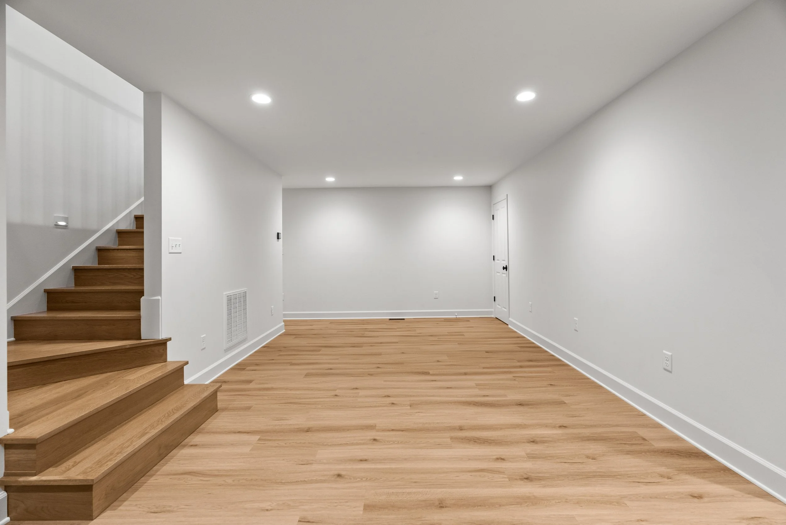 Empty room with wooden flooring, white walls, a staircase on the left, and a white door on the right.