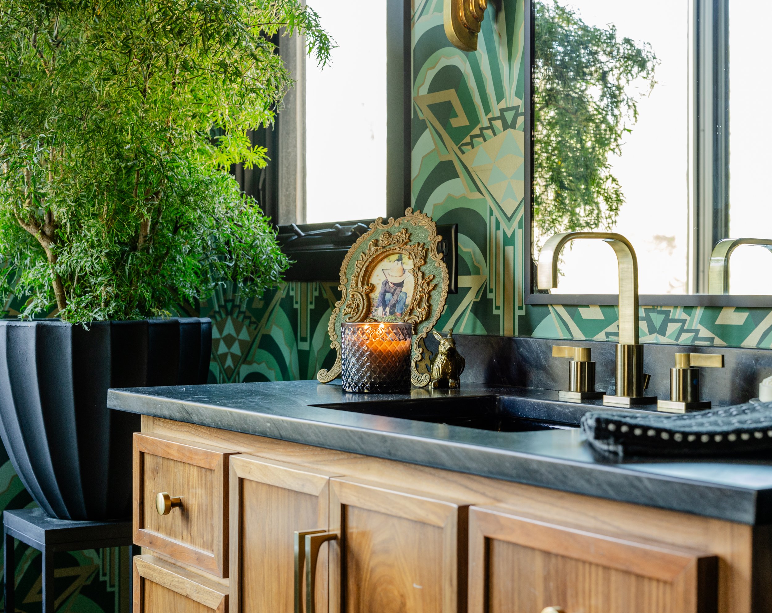 A kitchen countertop with a gold faucet, a decorative framed picture, a candle, and a small gold animal figurine, with a large green leafy plant on the side and a patterned wall behind.
