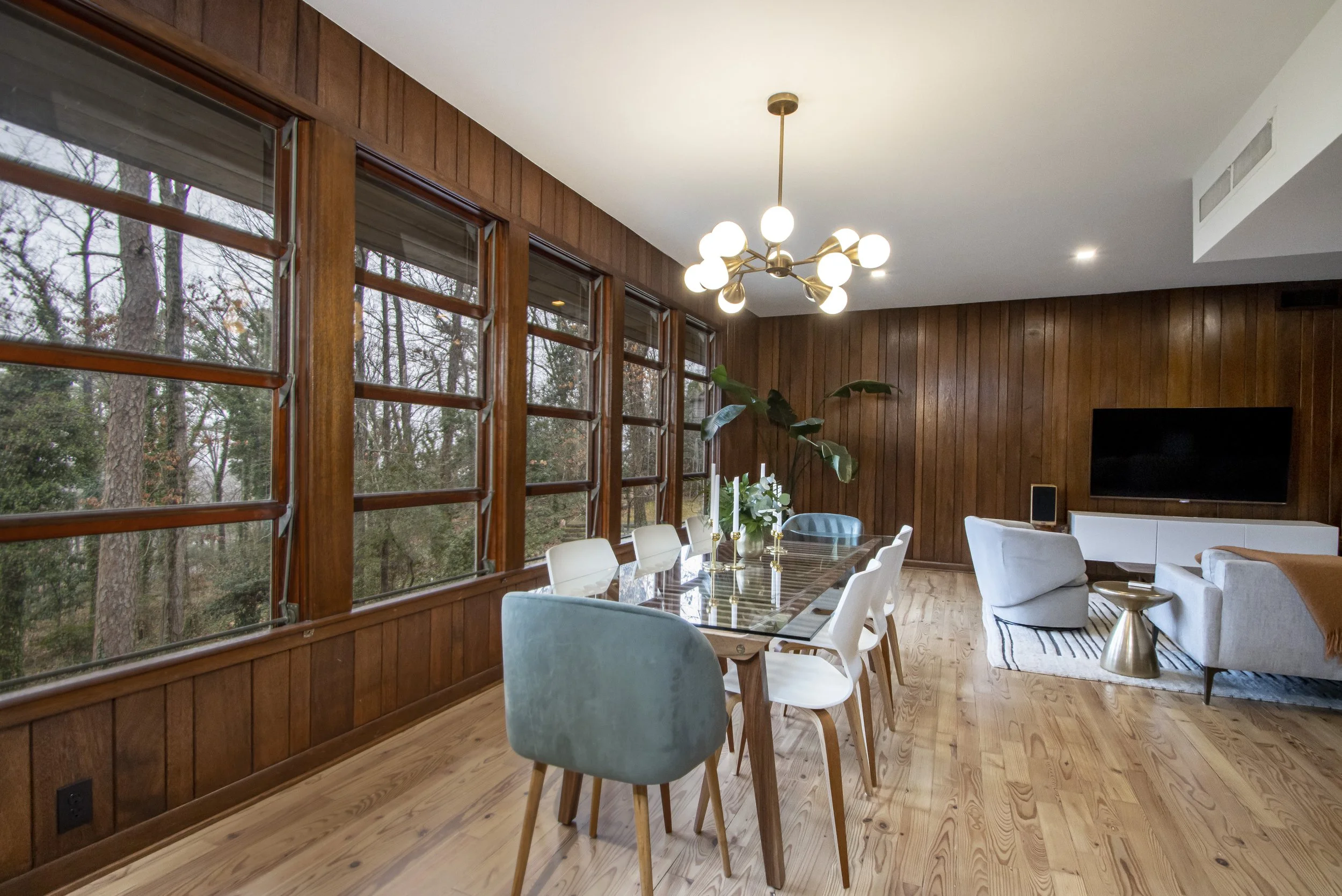 A dining and living room area with large windows showing trees outside, wooden wall paneling, a modern chandelier, a glass dining table with white and teal chairs, houseplants, a TV, and comfortable seating.