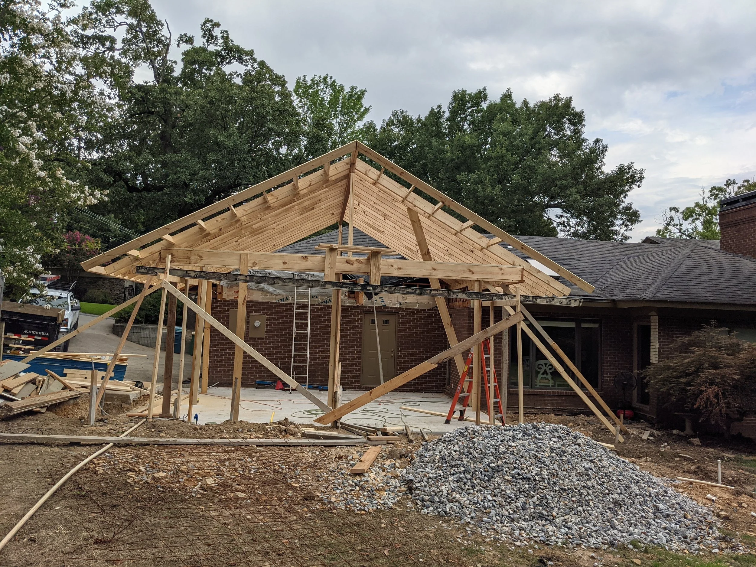 Construction site with a partially built roof structure on a brick house, trees in the background, and construction materials and debris on the ground.