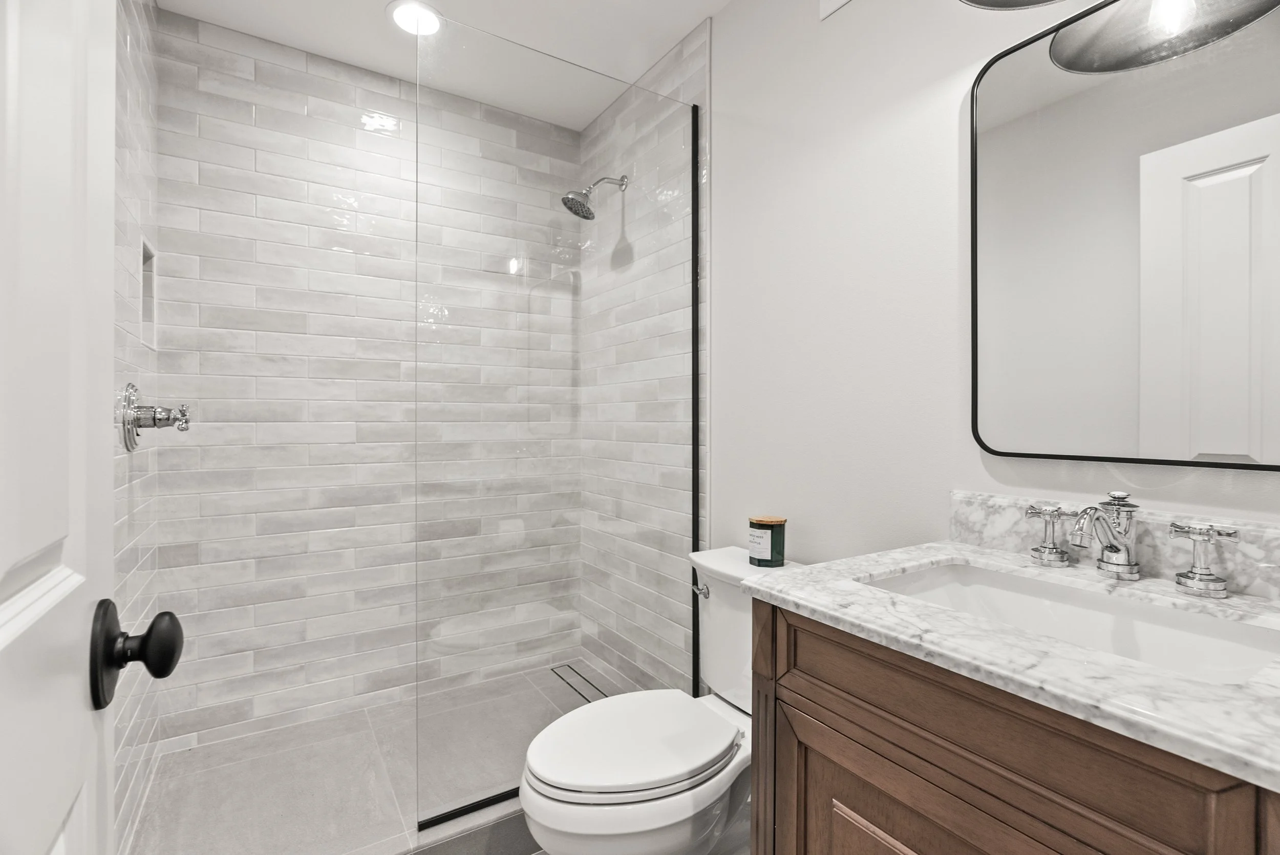 A modern bathroom with a walk-in shower with a glass divider, white subway tile walls, and a ceiling light. There is a toilet, a marble countertop with a wooden cabinet, a rectangular mirror, and a silver faucet. A candle or container is on the count
