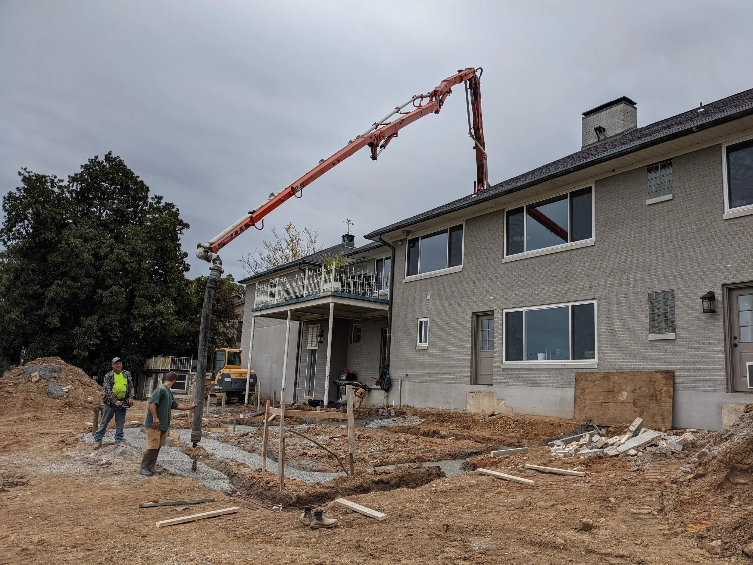 Construction workers working on a new sidewalk in front of a residential building with a concrete pump truck extending its arm to pour concrete.