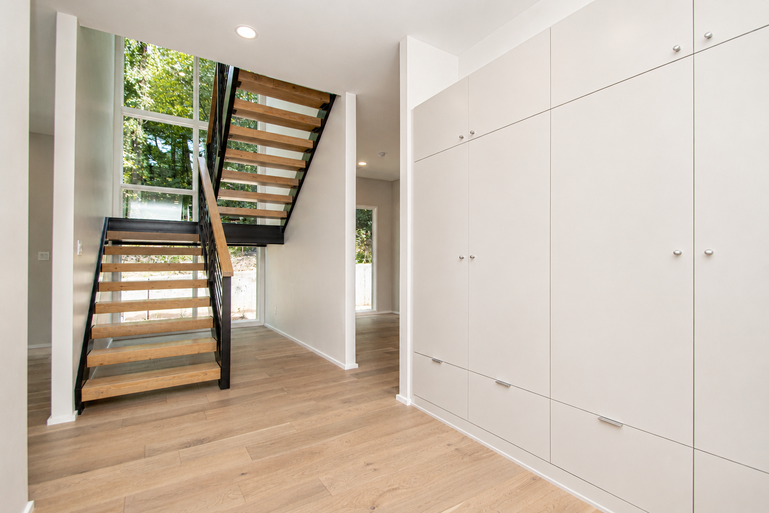 Interior of a modern home with light wooden floors, white walls, built-in white cabinets, a staircase with open risers and wooden treads, large windows showing green trees outside, and small recessed ceiling lights.