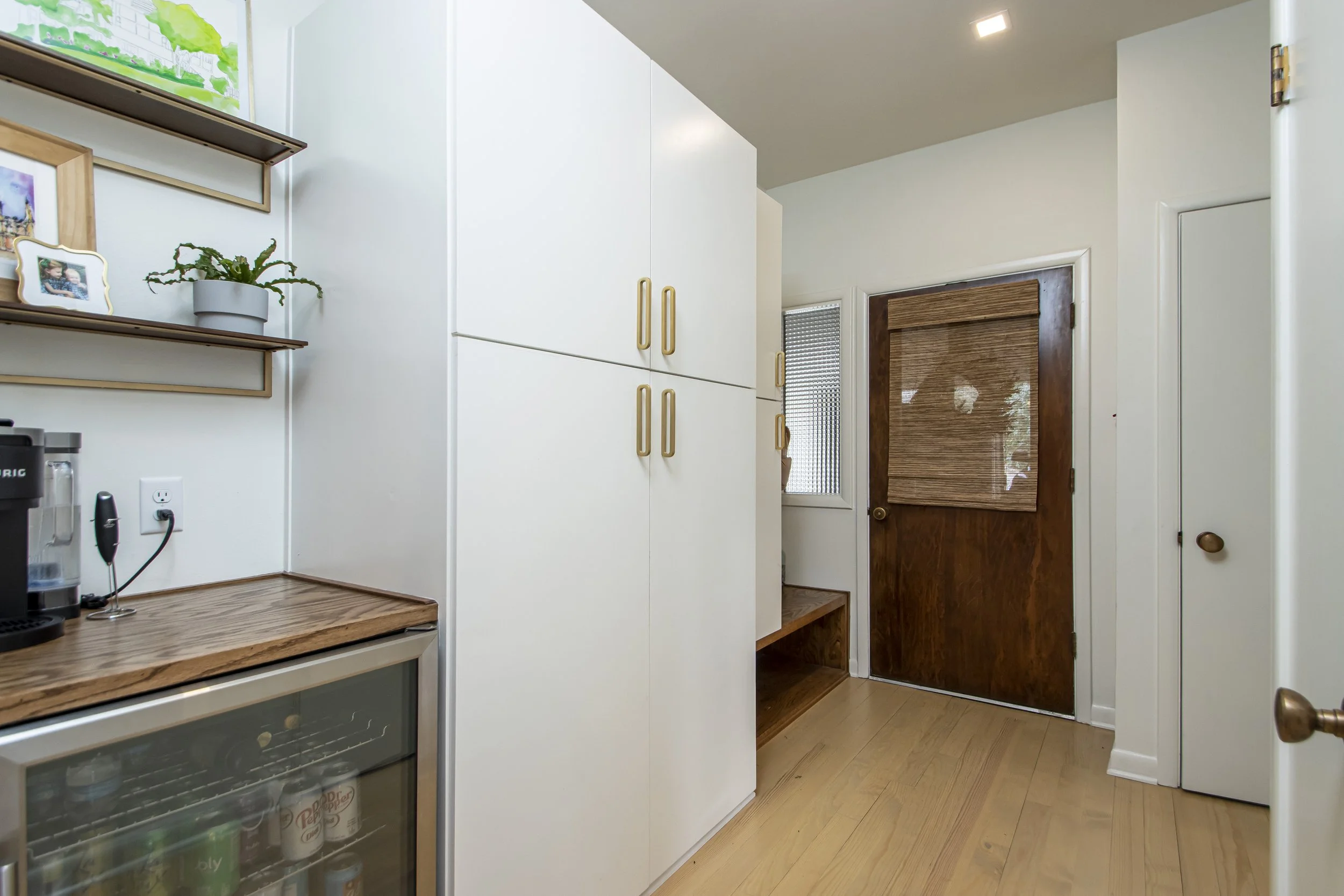A kitchen corner with white cabinets, a wooden countertop, a mini fridge with drinks, a coffee machine, and a wooden door with a woven shade, with a small window next to it.