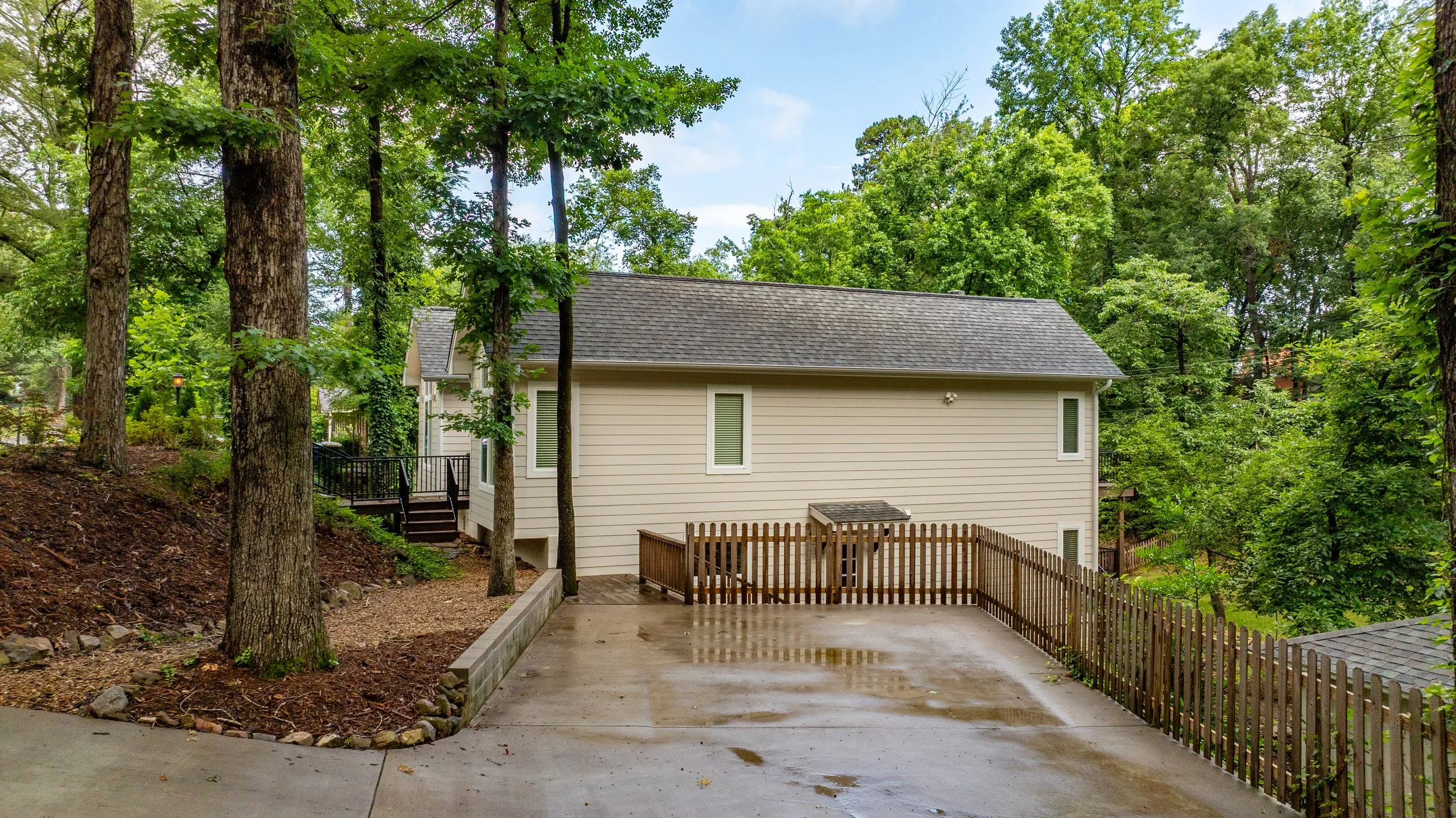 A backyard with tall trees surrounding a white house with a gray shingled roof, a wooden deck, and a fenced area. The ground is wet, indicating recent rain.